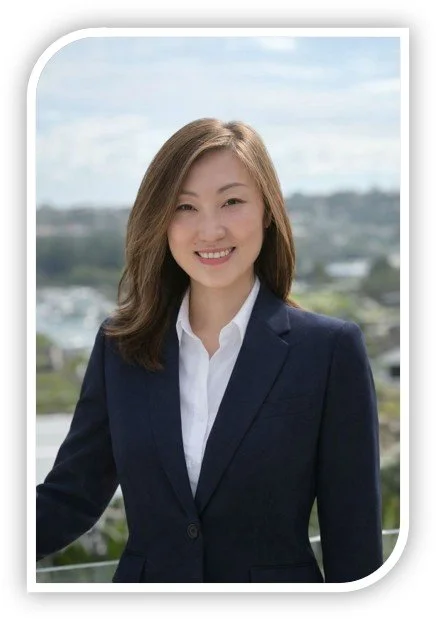 A woman with shoulder-length brown hair wearing a navy blazer and white shirt, smiling outdoors with a cityscape and cloudy sky in the background.