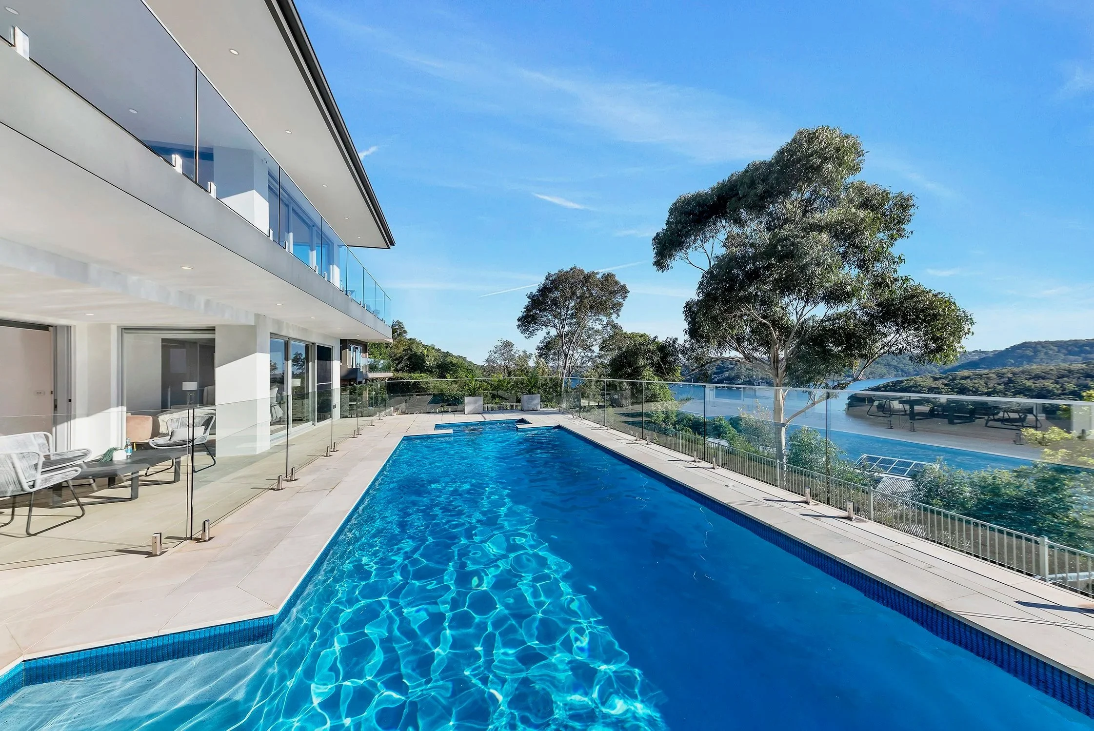 Modern house with a large swimming pool outside, glass balcony railing, and scenic view of trees and hills under a clear blue sky.