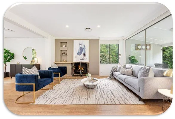 Modern living room with a gray sofa, a blue armchair with gold frame, a fireplace, a white rug, and large windows showing greenery outside.
