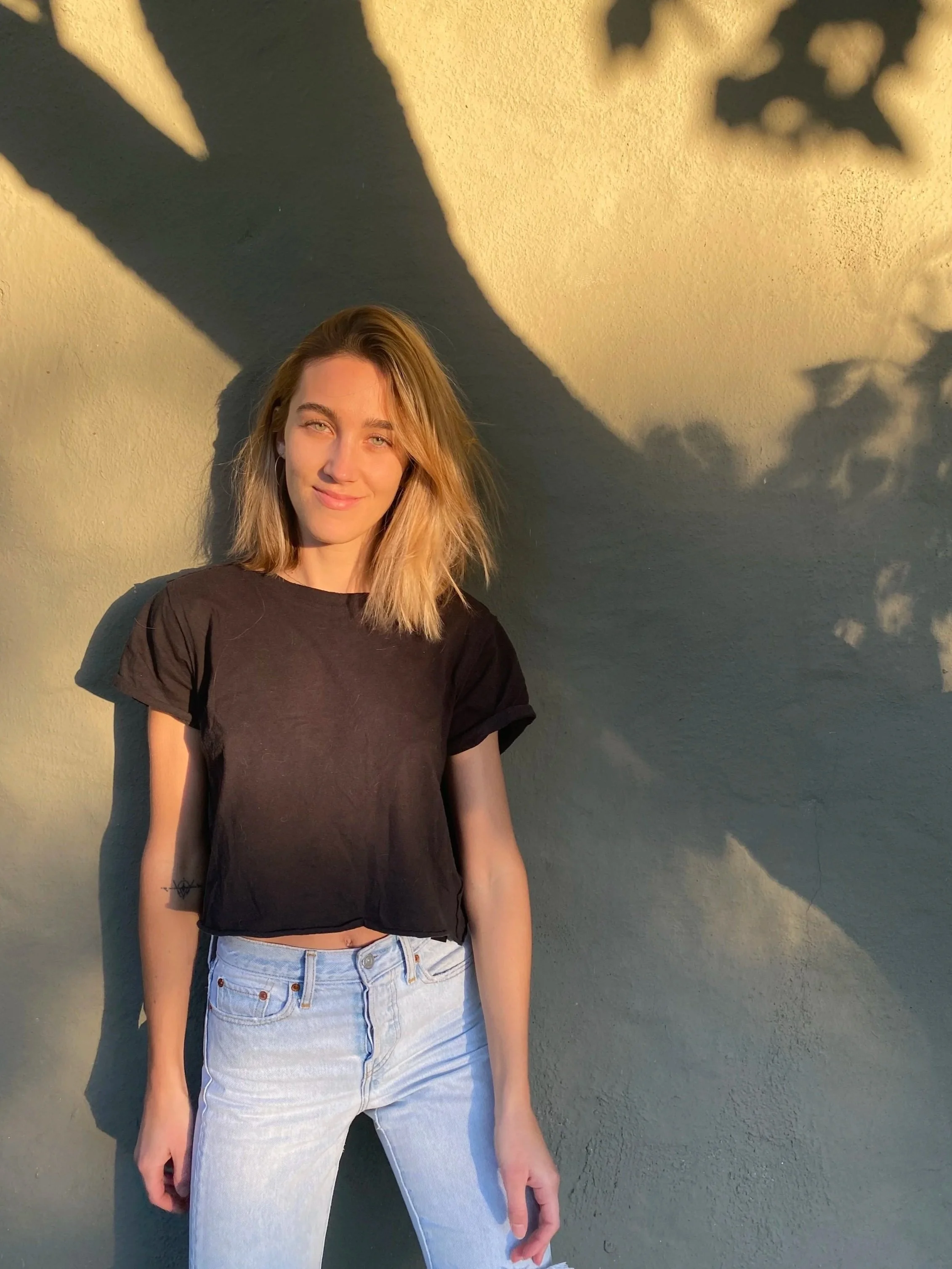 A young woman with blonde hair, wearing a black t-shirt and light blue jeans, standing against a wall with shadows of leaves cast on it, illuminated by sunlight.