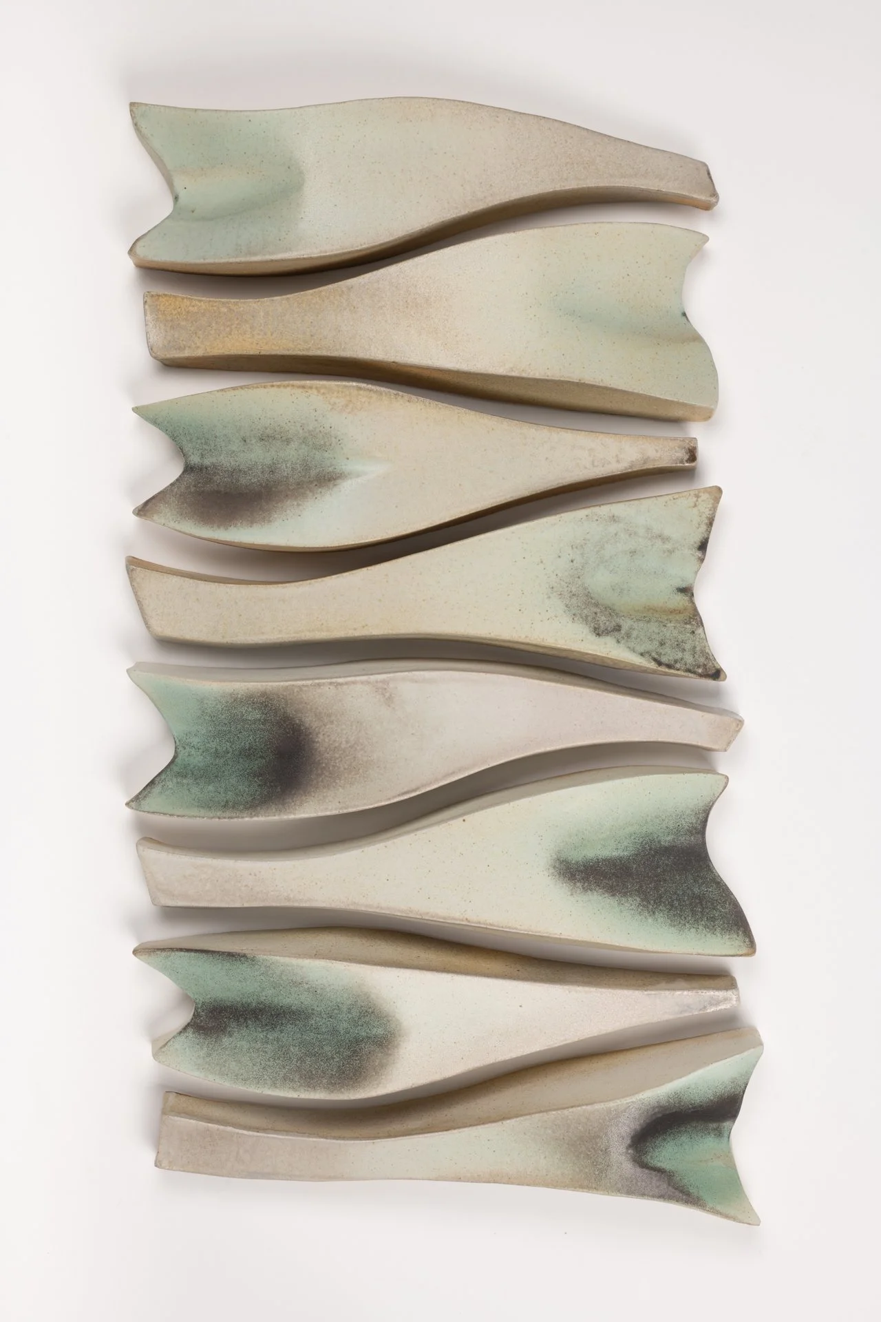 A wall installation of eight ceramic fish-shaped abstract objects arranged vertically, with muted beige, green, and black speckled  colors and textured surfaces. 