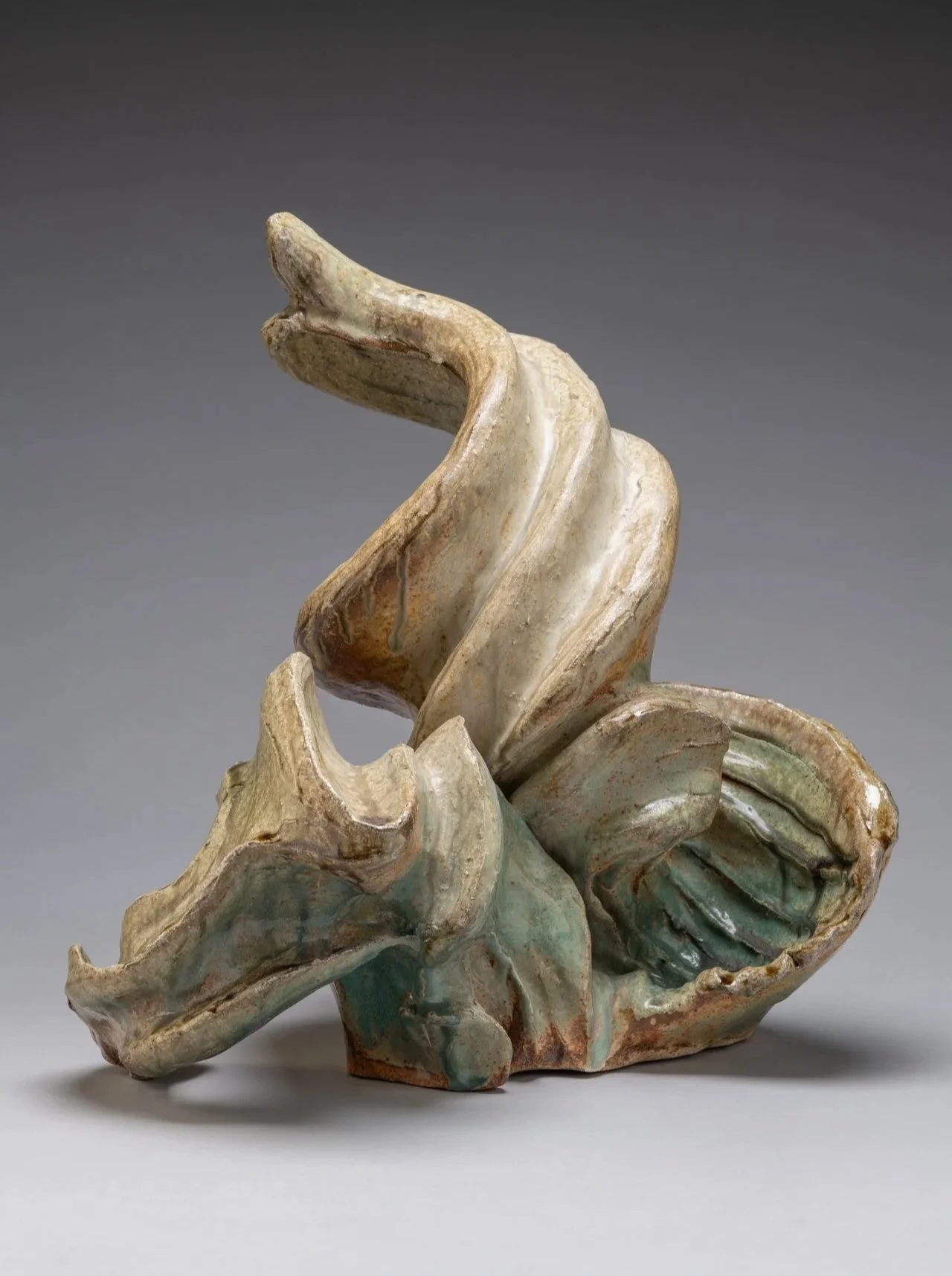 An abstract ceramic sculpture resembling a stylized wave with flowing curvilinear textured surfaces, and a mix of beige, brown, and greenish hues.