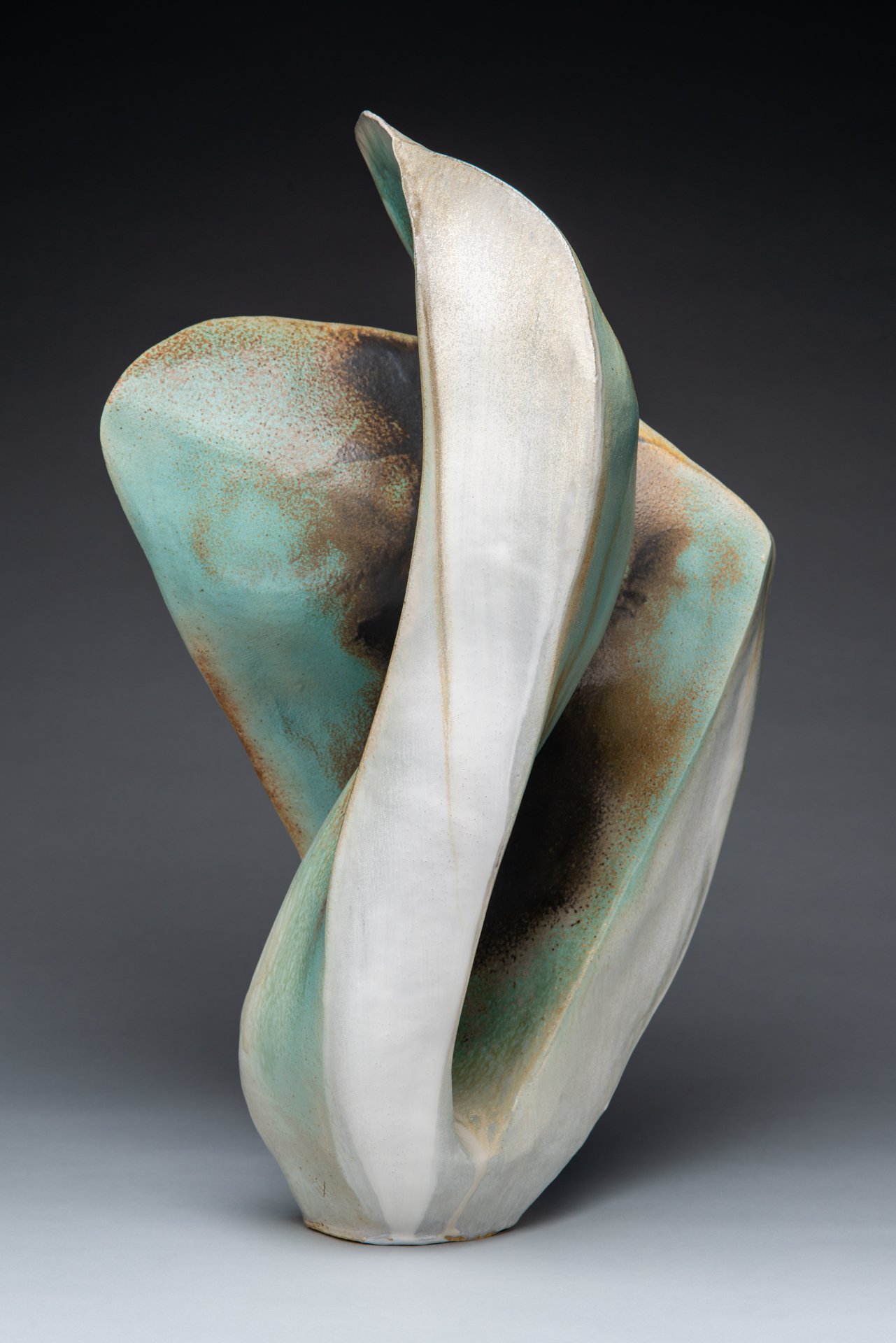 Abstract ceramic sculpture with curved, overlapping surfaces in shades of white, green, and brown against a dark background.