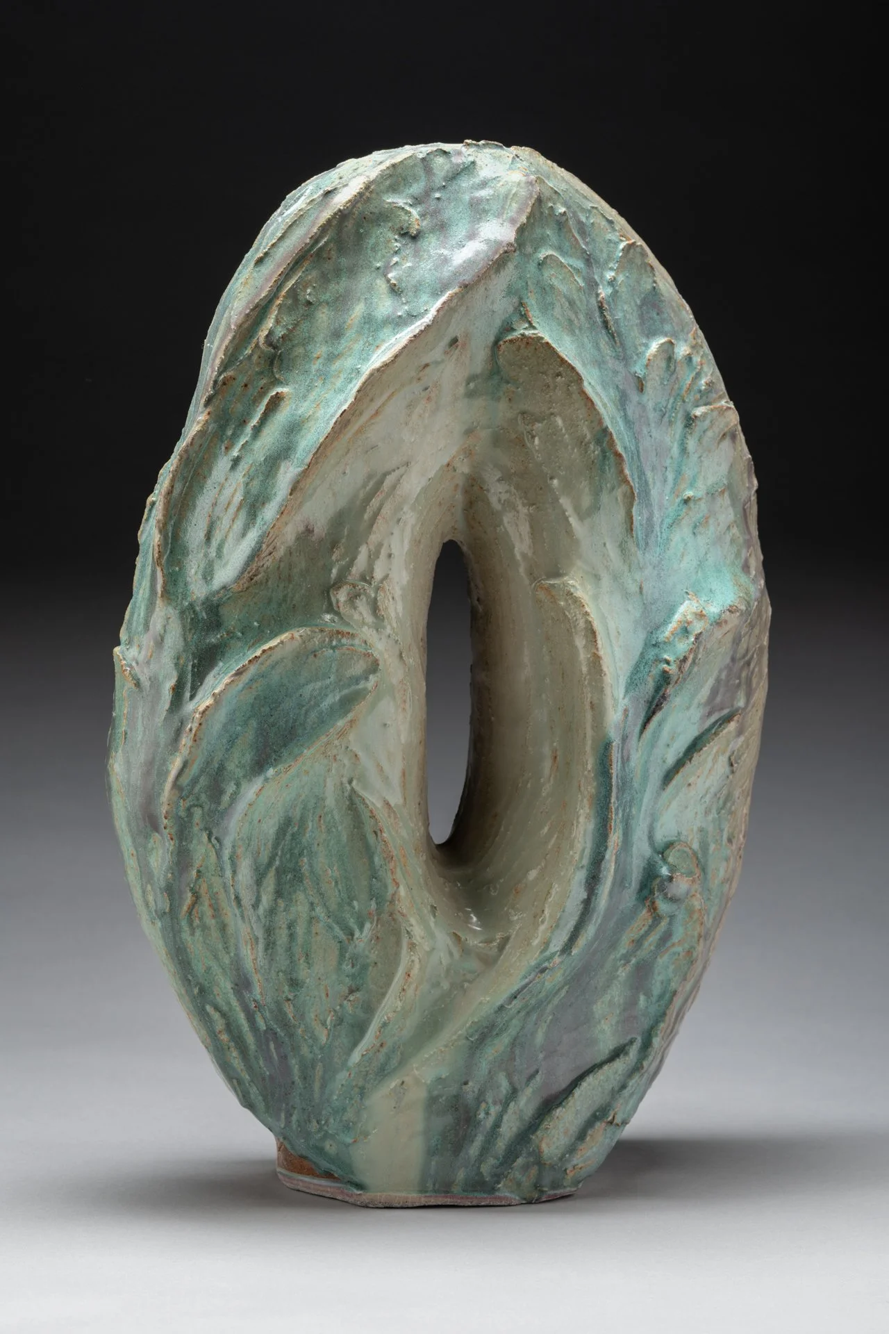 An oval ceramic sculpture with a hole in the center, styled with textures like waves in swirling green, white, and blue glazes.