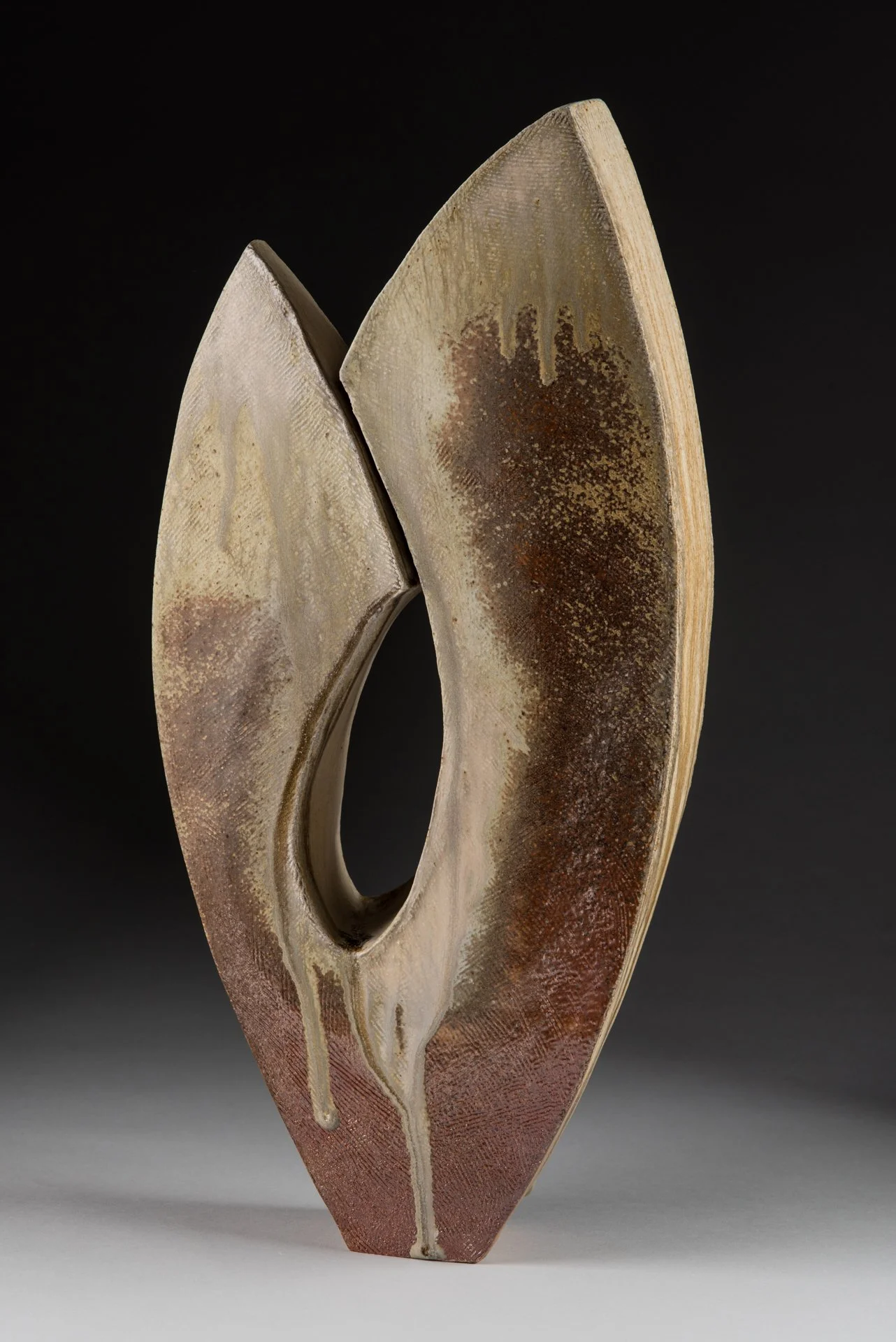 A modern ceramic sculpture with curved, crescent shapes and a textured surface, featuring earthy tones and drips of glaze against a black background.