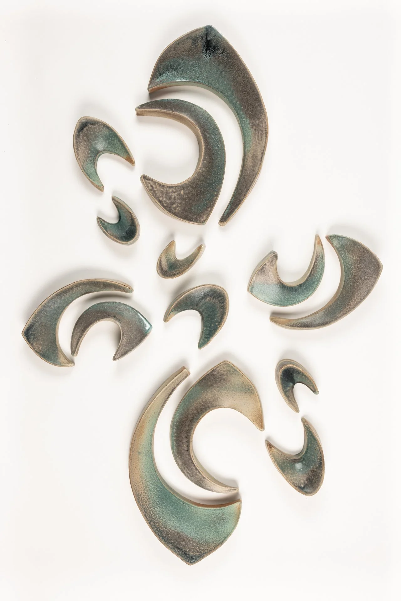 Wall installation of decorative ceramic crescent-shaped pieces in various sizes and shades of green, brown, and beige on a white background.