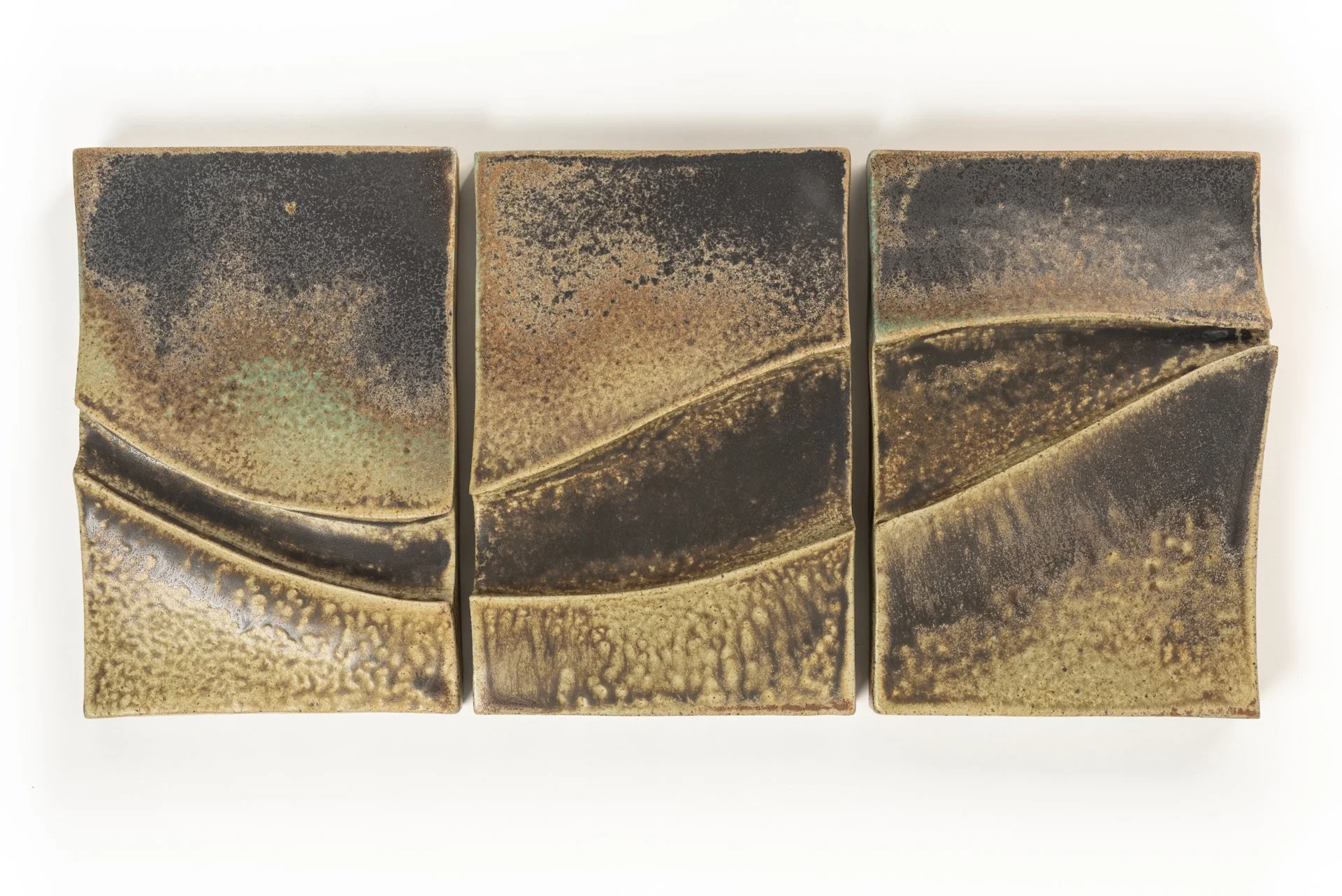 Three rectangular ceramic pieces with a textured, earthy glaze in shades of brown, black, and green, arranged side by side on a white surface.