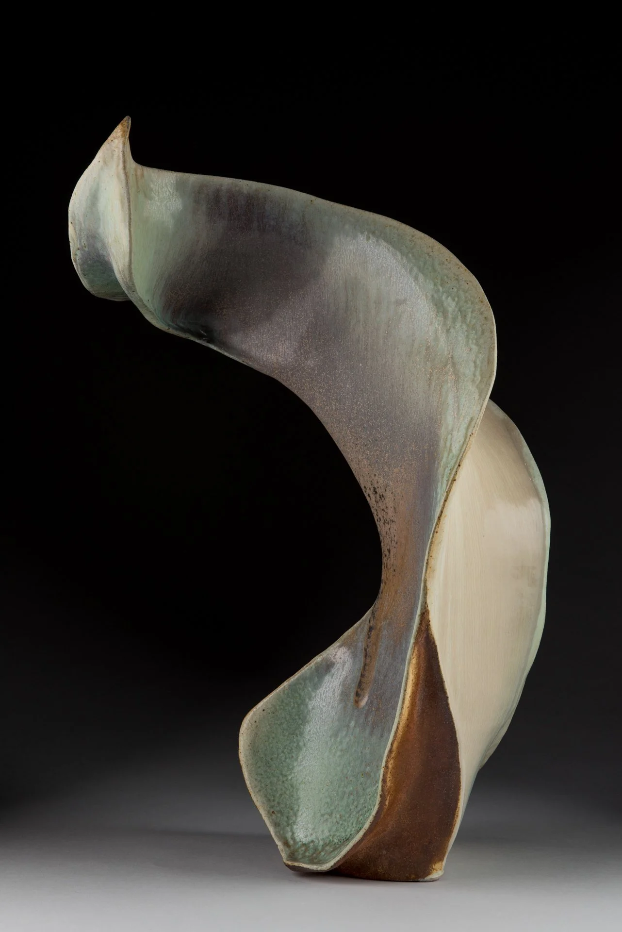 Abstract ceramic sculpture with flowing curves and earth-tone glazes against a black background.