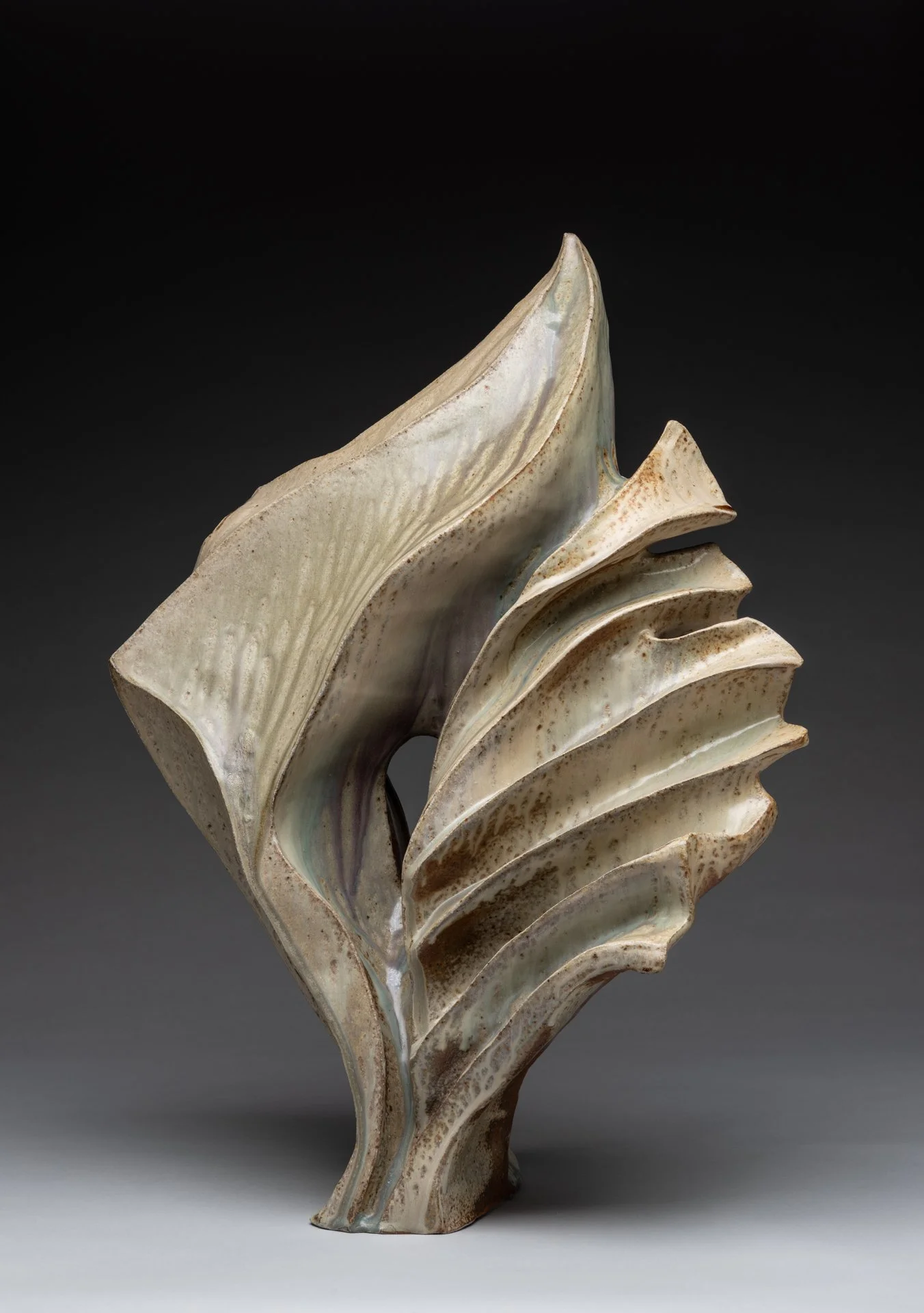 Abstract ceramic sculpture resembling a stylized leaf with layered, curved forms in earth tones against a black background.