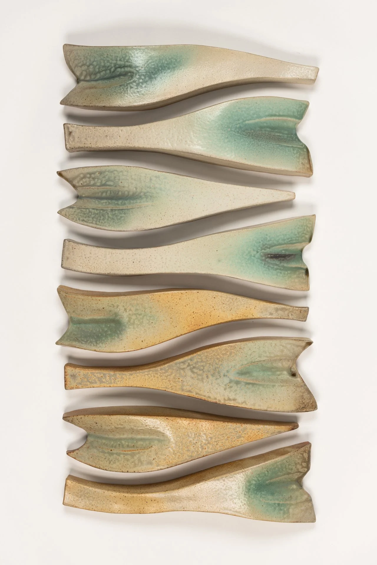 A wall installation of eight ceramic fish-shaped abstract objects arranged vertically, with muted yellow, white and green colors and textured surfaces.