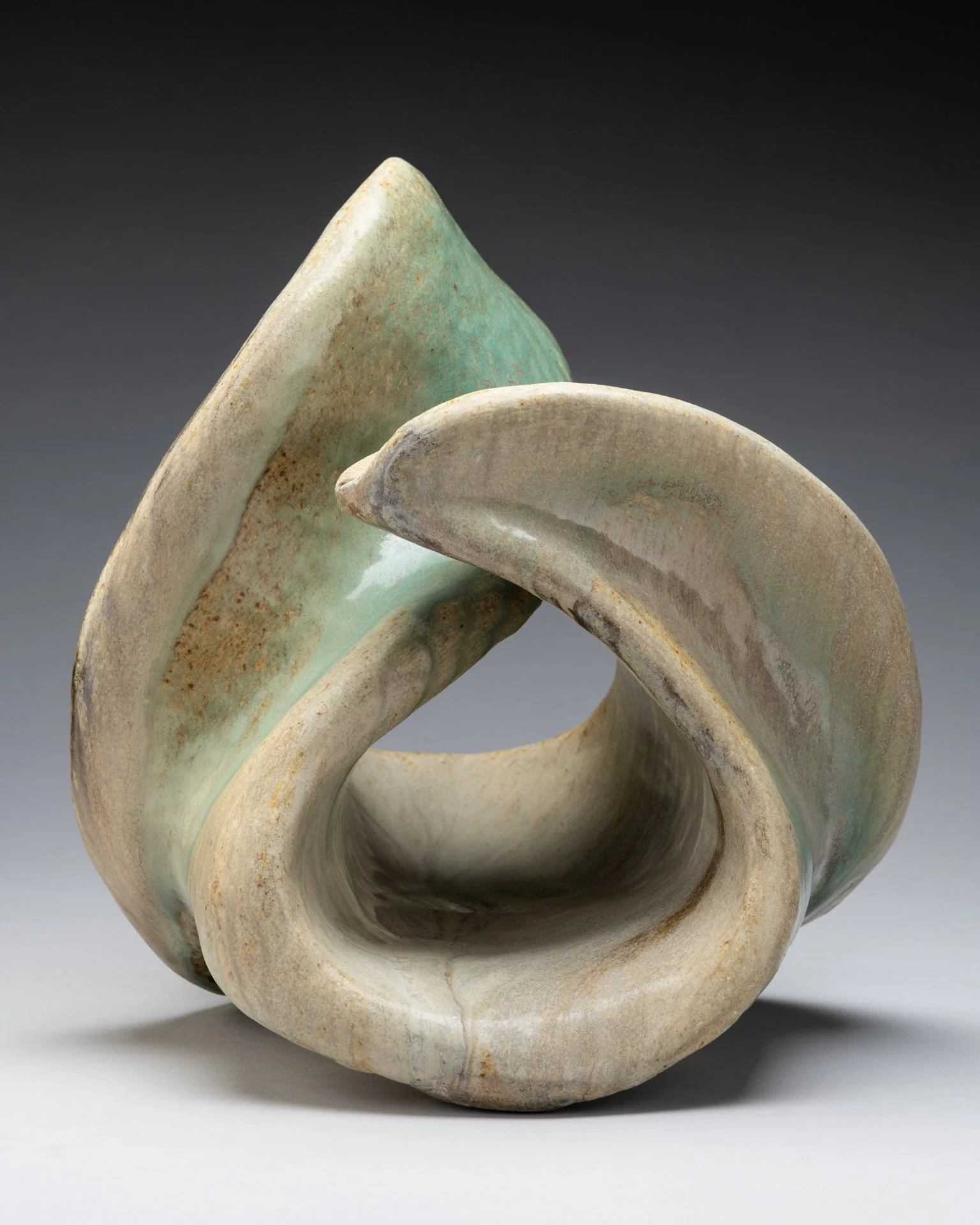 A gently rounded ceramic sculpture with a smooth surface, featuring shades of pale green and beige, displayed against a dark gray background.