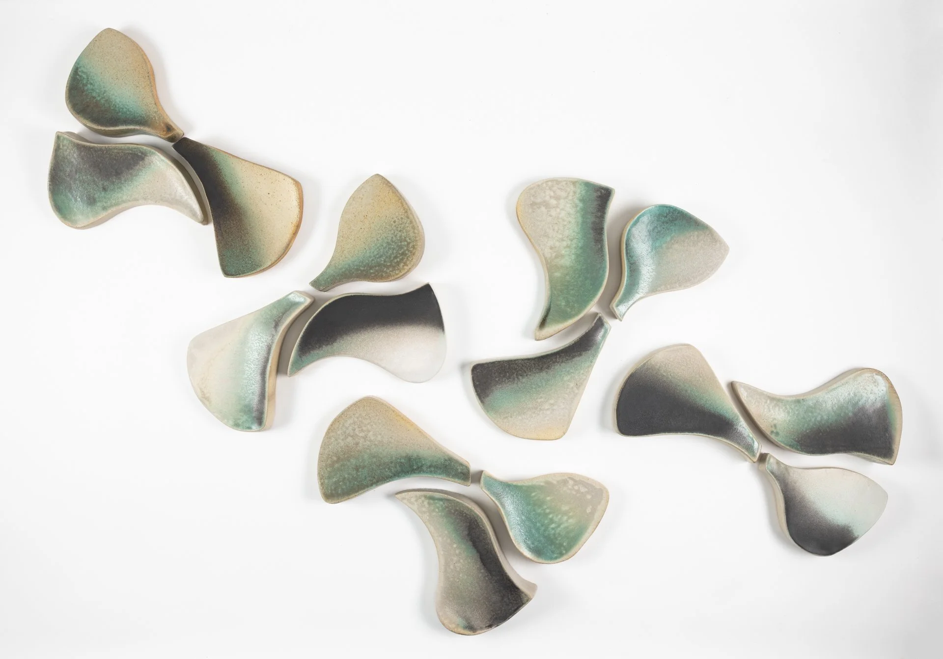 A collection of ceramic pieces shaped like flower petals with a gradient glaze in green, beige, black, and cream colors arranged on a white wall.