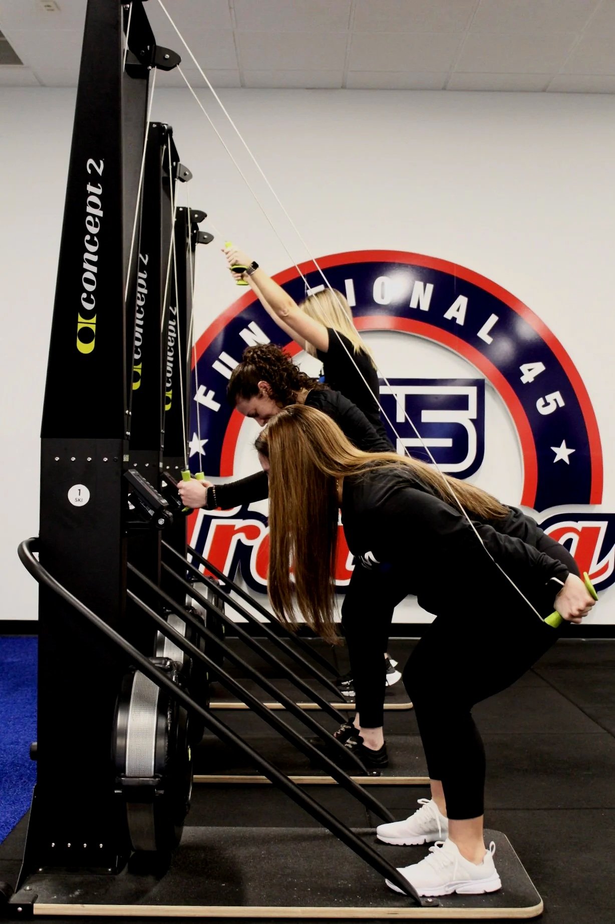 Three women using a vertical climbing machine at the gym with a fitness-related logo on the wall behind them.