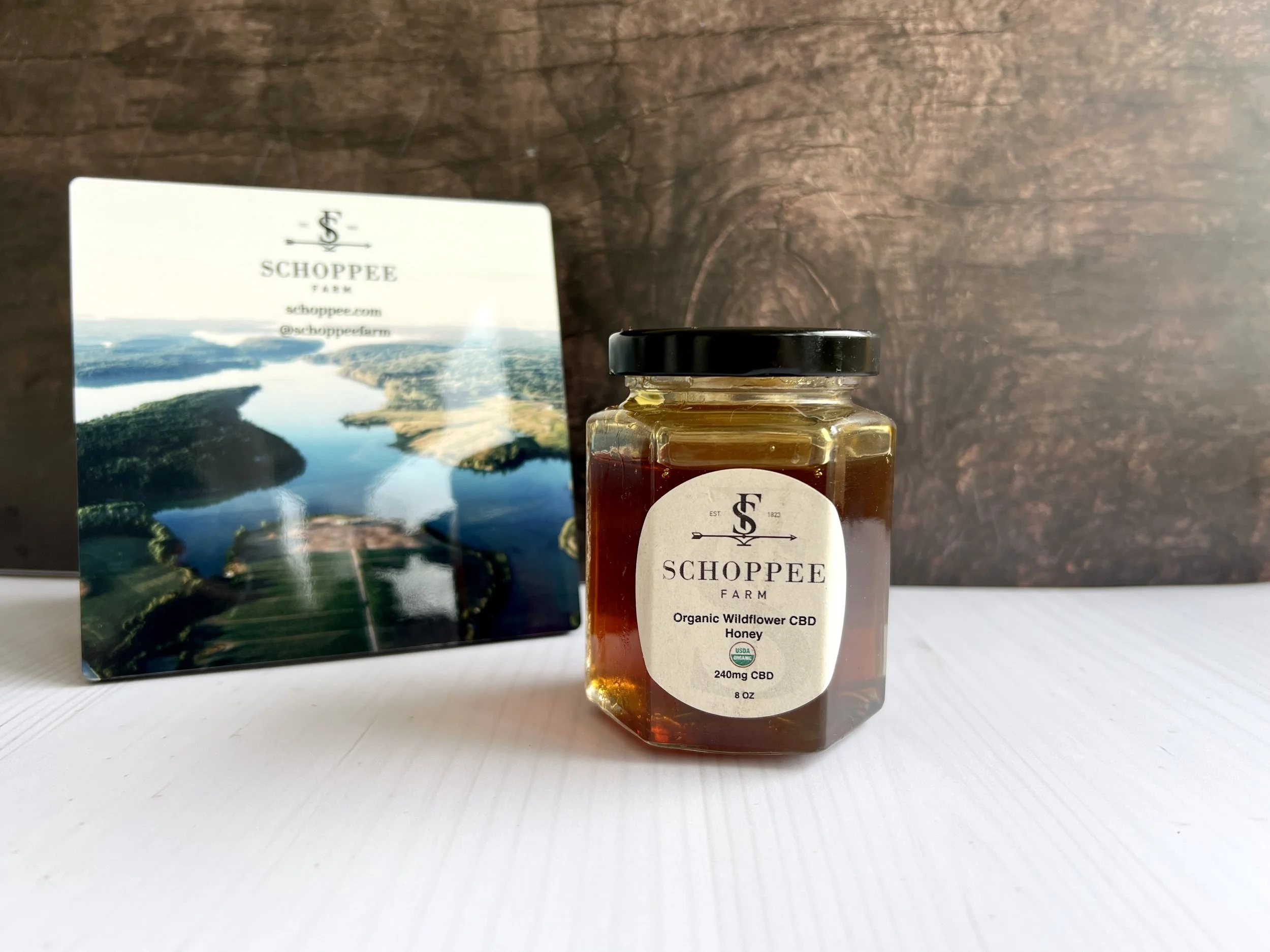A jar of Schoppée Farm Organic Wildflower CBD Honey with a black lid, placed on a white surface, with a rectangular card featuring an aerial view of a river landscape in the background.