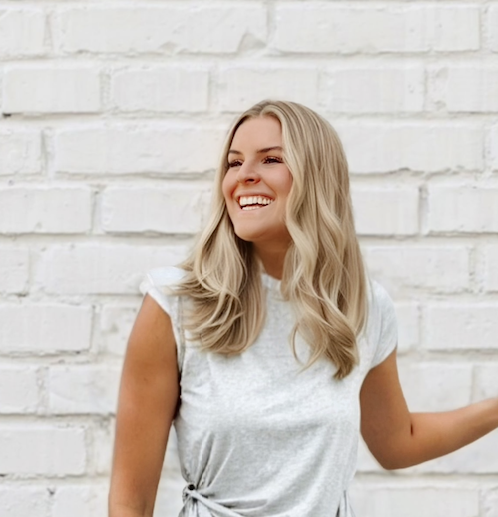 A smiling young woman with long blonde hair standing outdoors in front of a white brick wall.