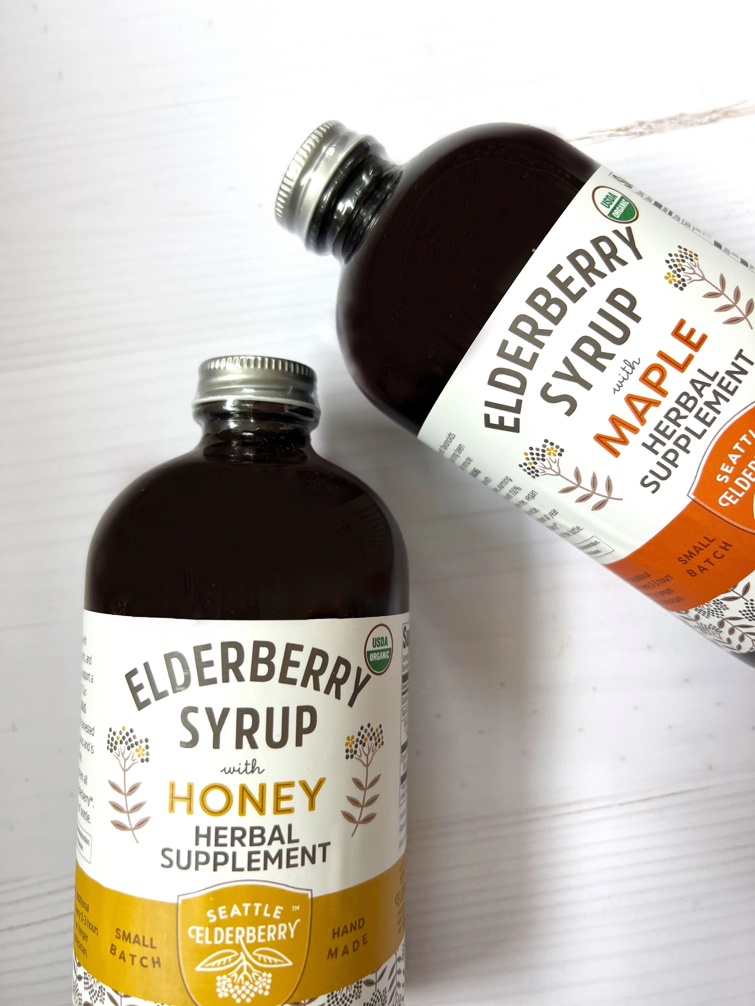 Two bottles of Elderberry Syrup sitting on a white surface, one labeled Elderberry Syrup with Honey and the other Elderberry Syrup with Maple, both indicating herbal supplements for health.