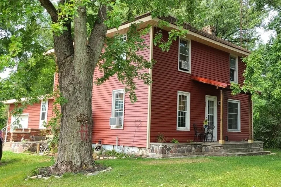 Red two-story house with white trim, surrounded by green trees and lawn. Front porch with small table and chairs, stone steps leading to the entrance.