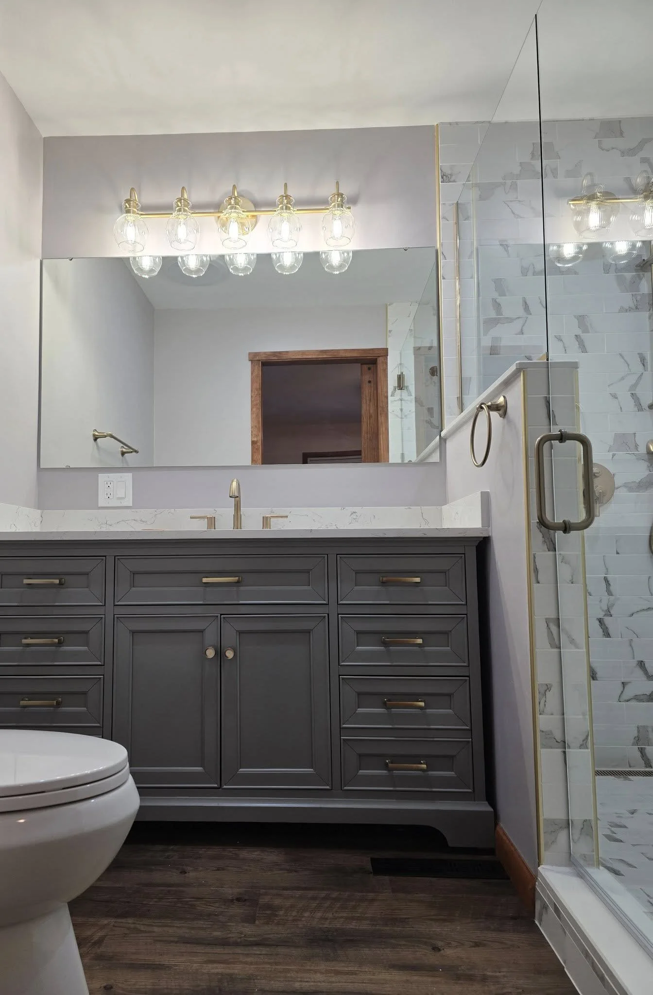 A modern bathroom with a gray vanity, marble countertop, large mirror, and a glass-enclosed walk-in shower with marble tile walls, a wooden door, and a wood floor.