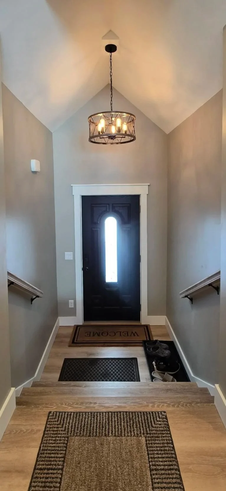 Entrance hallway with a black front door featuring a tall, narrow window, a decorative chandelier hanging from the ceiling, a welcome mat with 'WELCOME' written on it, and shoes placed on a shoe tray near the stairs, with beige walls and hardwood flooring.