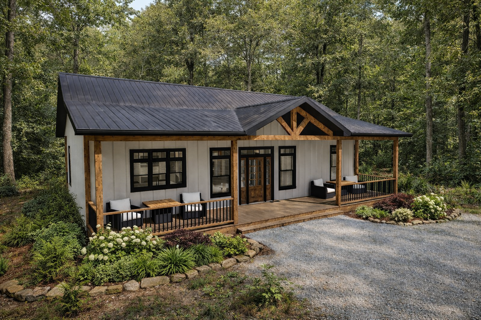 A cozy house with a black metal roof, white siding, and a front porch with seating, set in a wooded area with landscaped flower beds and a gravel driveway.