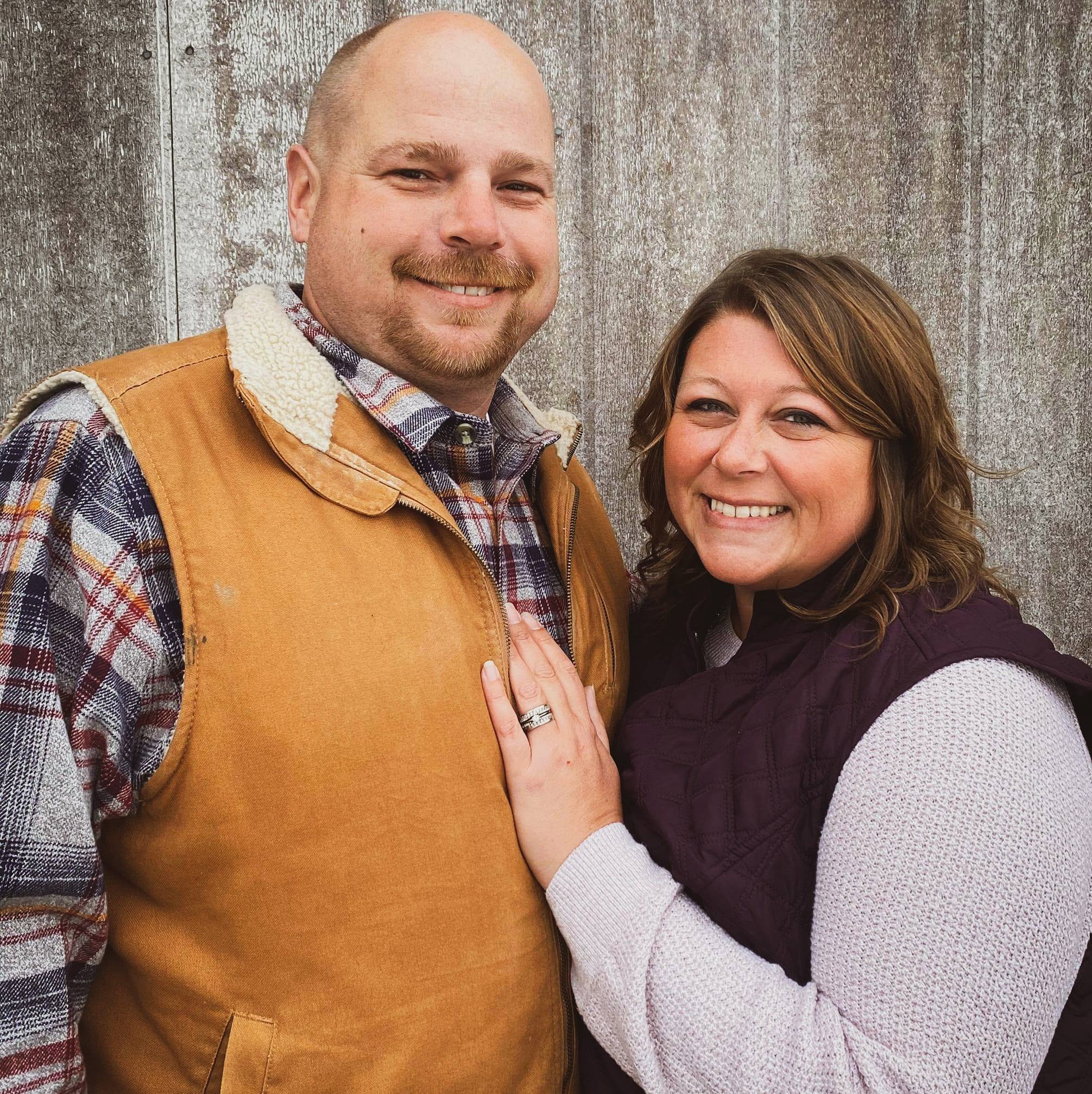 A smiling man and woman standing close together outdoors, with a weathered wooden wall in the background. The man is wearing a plaid shirt and a tan vest with a sherpa collar, and the woman is wearing a white sweater with a purple quilted vest. The woman has her hand on the man's chest, showing a ring on her finger.