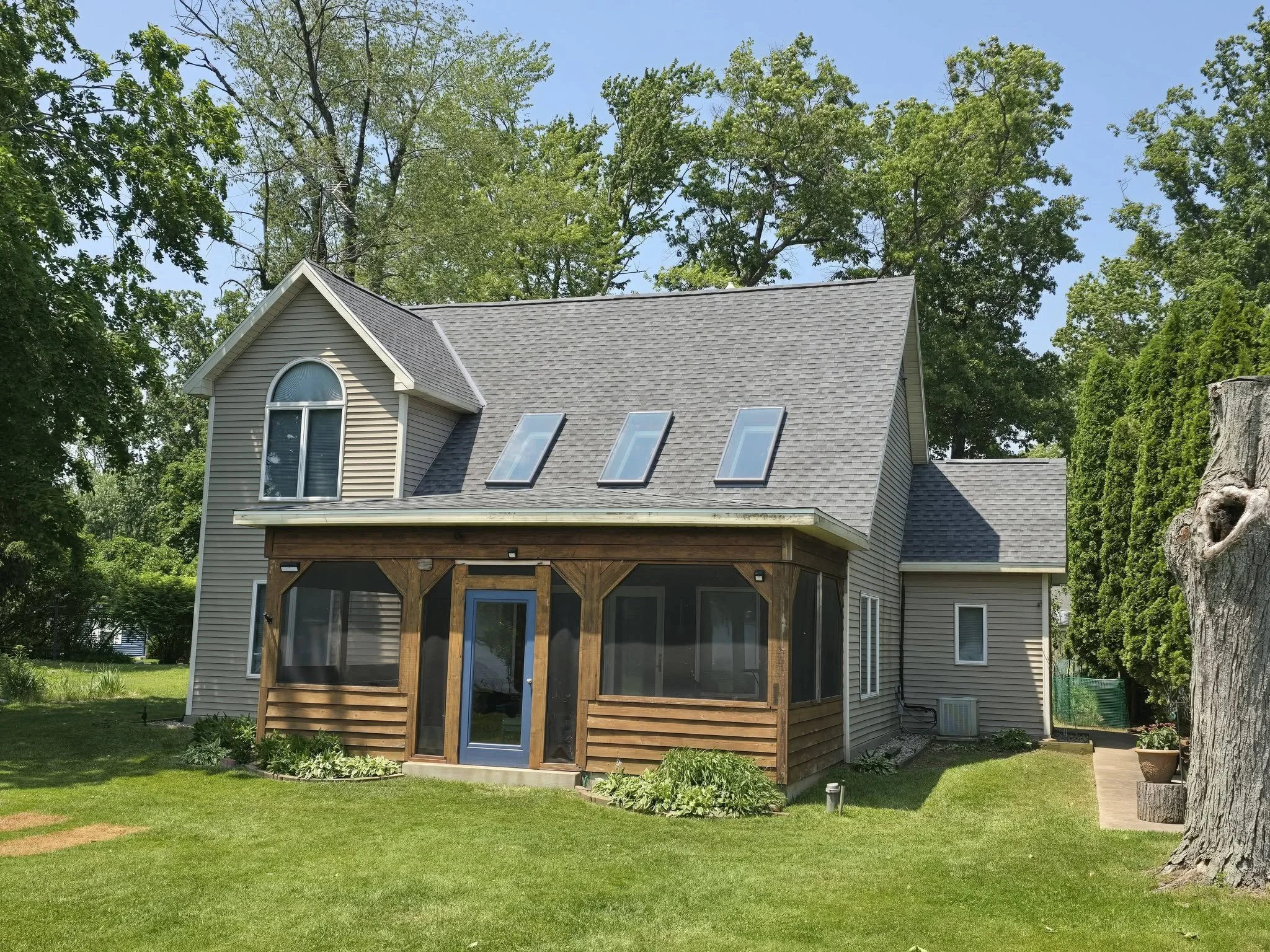 A two-story house with beige siding, a gabled roof, three skylights, and a screened porch with wooden framing. The house is surrounded by green grass, trees, and shrubs, with a large tree trunk on the right side.