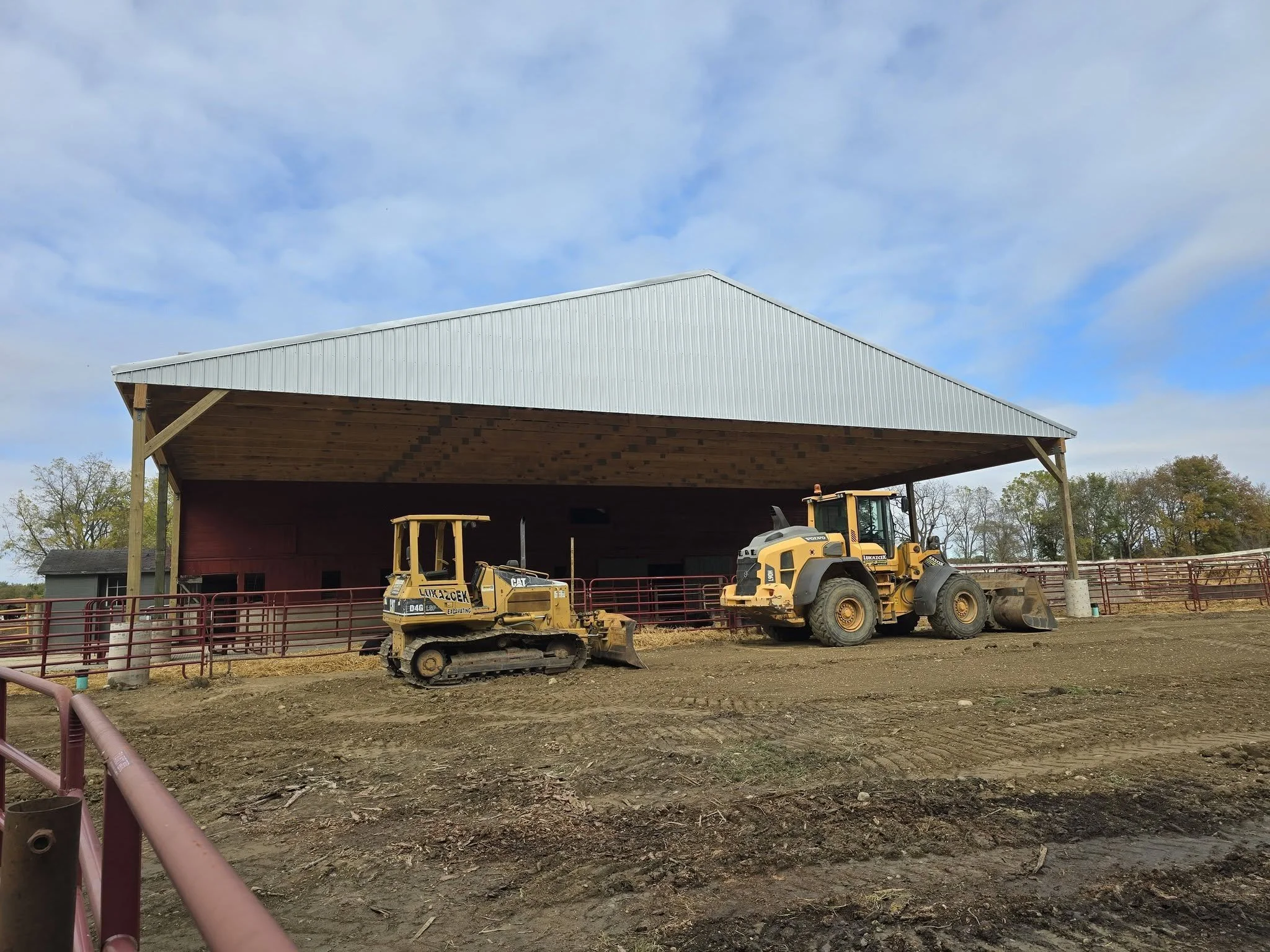 Construction site on a farm with two yellow construction vehicles in front of a large open barn with a metal roof and wooden interior, surrounded by a red metal fence and trees in the background.