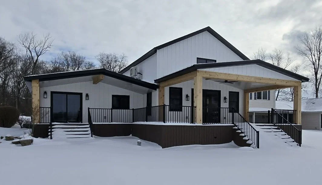 Modern two-story house with a large wooden porch, black metal railings, snow-covered ground, and leafless trees in the background.