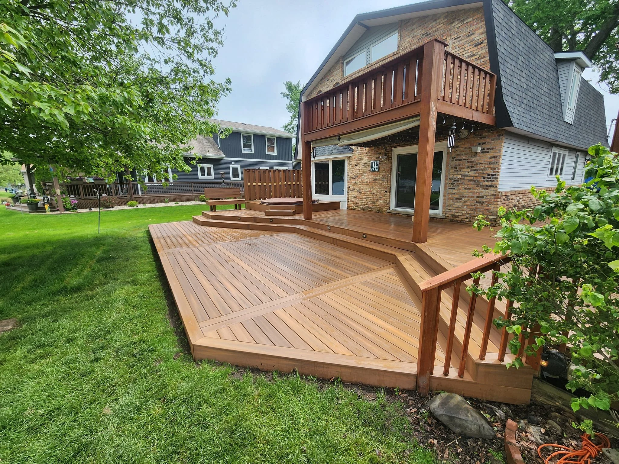 Backyard with wooden deck, stairs, and balcony of brick and siding house, green lawn, trees, and neighboring houses.