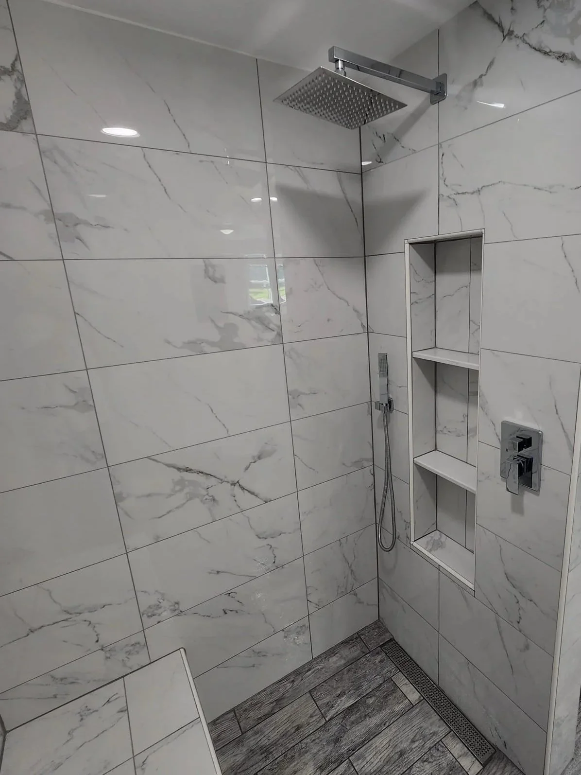 A modern walk-in shower with large marble-like tiles, a square rainfall showerhead, a built-in niche with shelves, a handheld shower, and a linear floor drain.