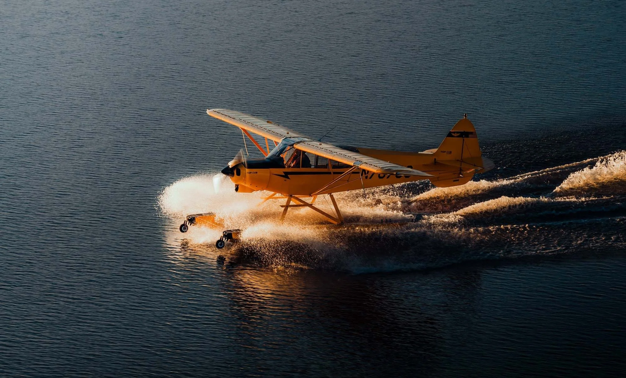 A yellow seaplane flying low over water, creating a splash as it skim the surface.