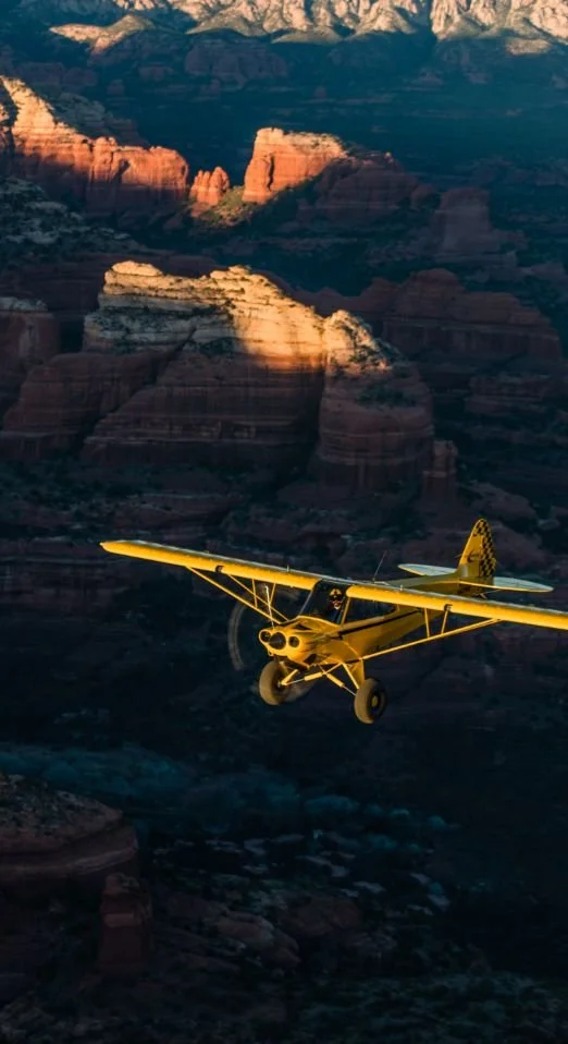 A yellow airplane flying over the Grand Canyon during sunset, with layered rock formations visible in the background.