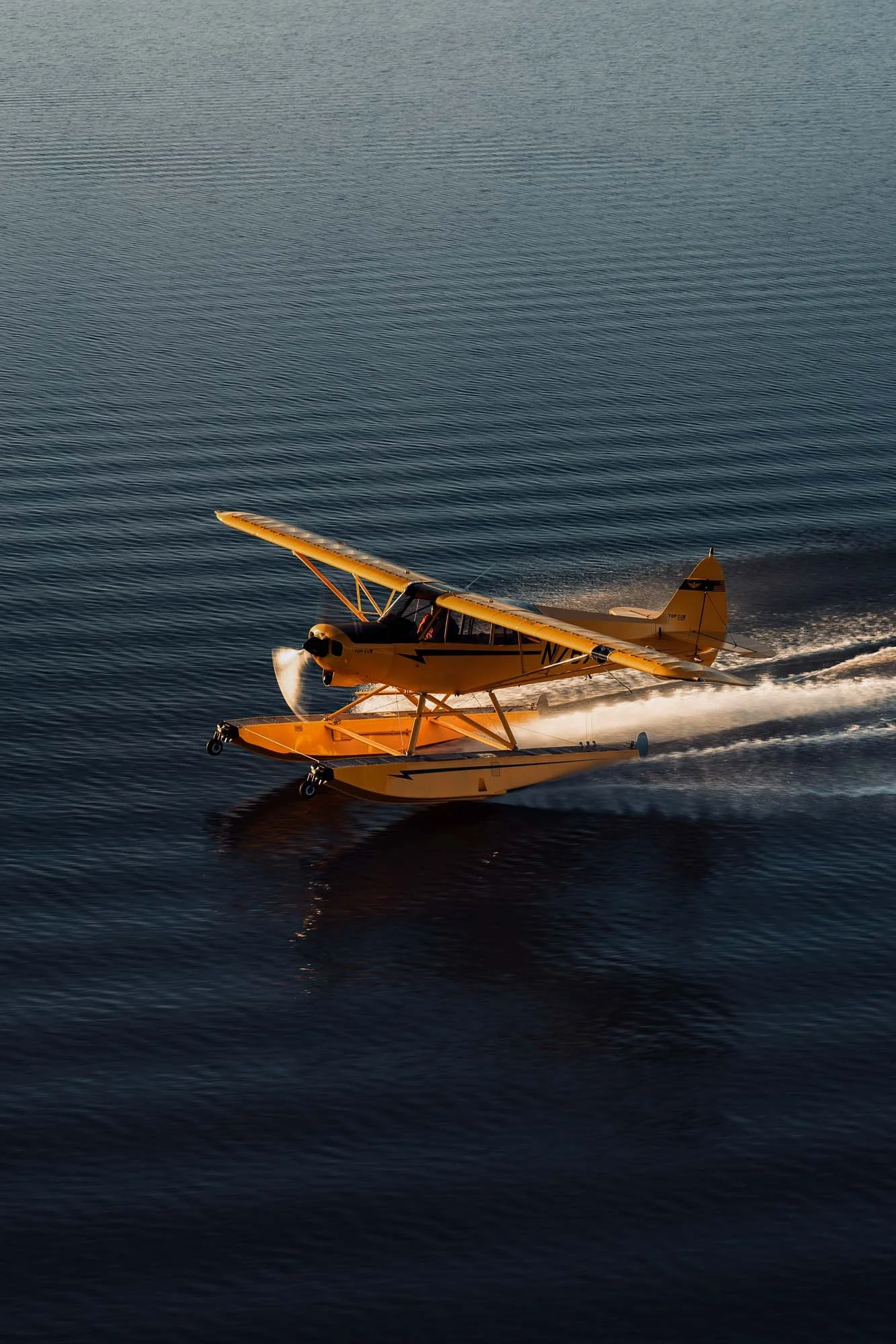A yellow seaplane flying low over a calm body of water, creating a wake behind it.