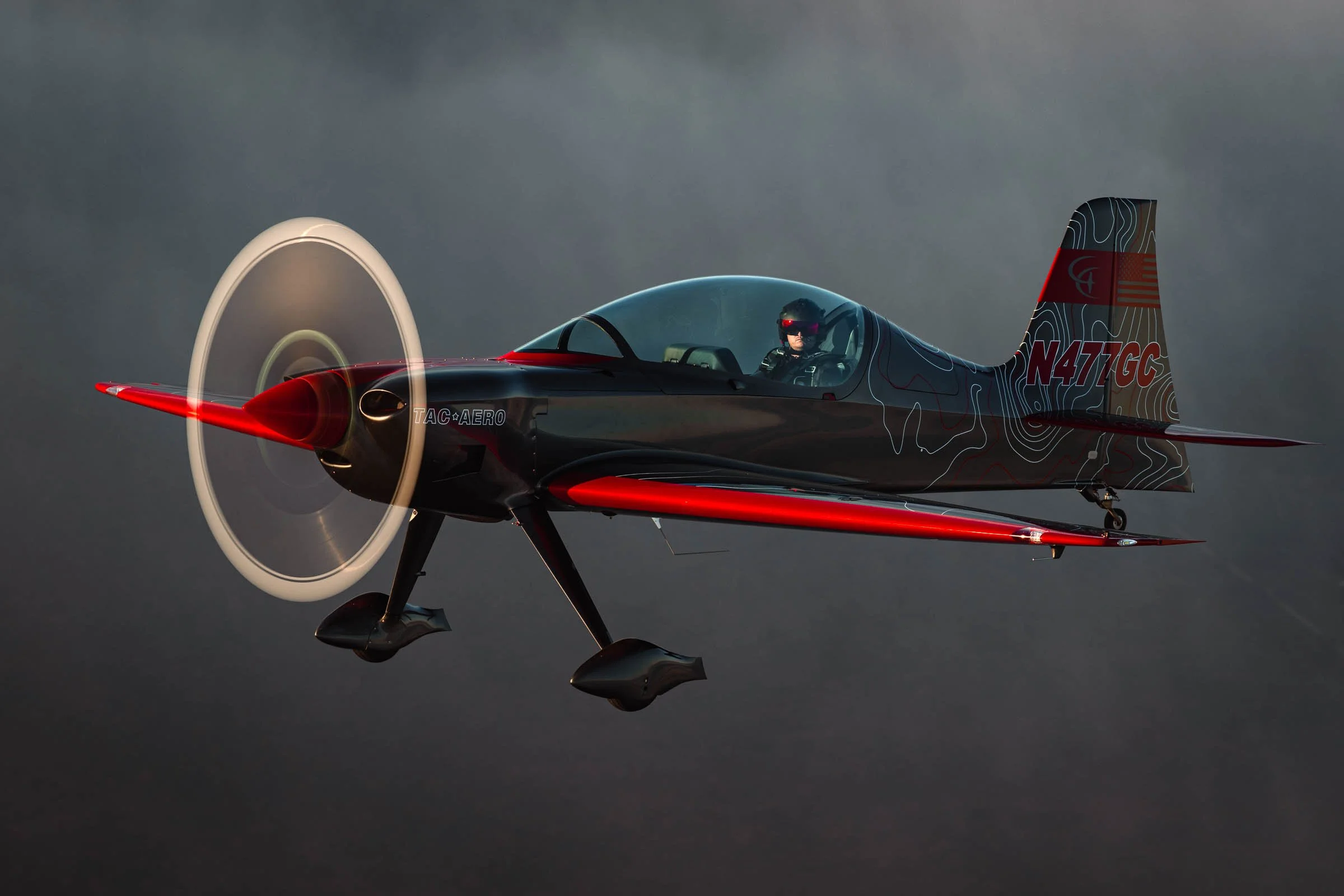A sleek black and red aerobatic aircraft flying against a dark sky, with a pilot wearing a helmet and headset visible through the canopy.
