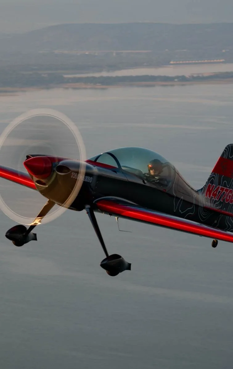A small propeller airplane flying over water with a distant shoreline and hills in the background.