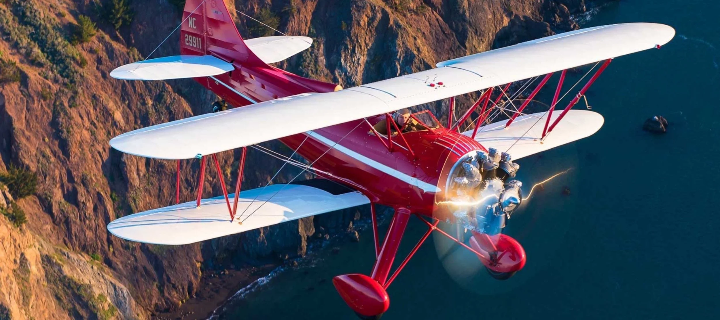 A red and white biplane flying over a rocky coastline near the ocean, with the engine producing sparks and lightning effects.