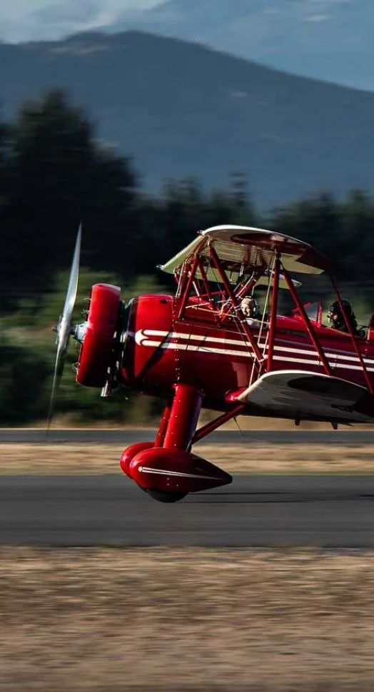A red vintage biplane taking off from a runway with mountains in the background.
