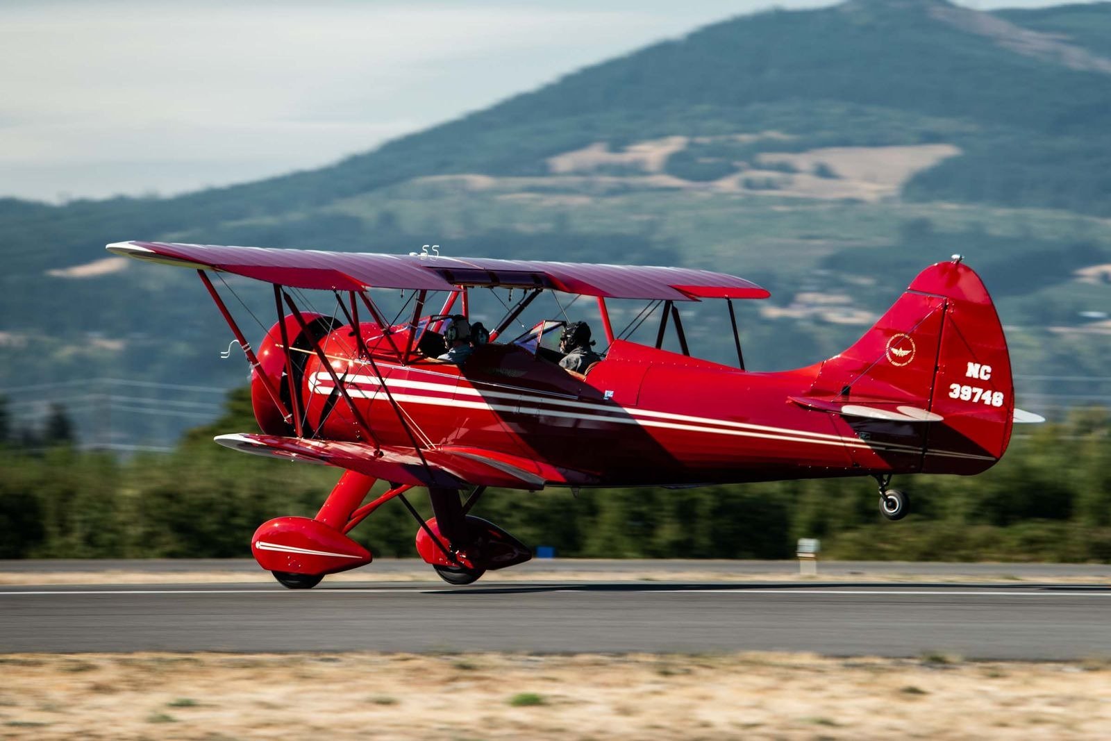 Red biplane airplane taking off from runway with two pilots inside, mountain landscape in background.