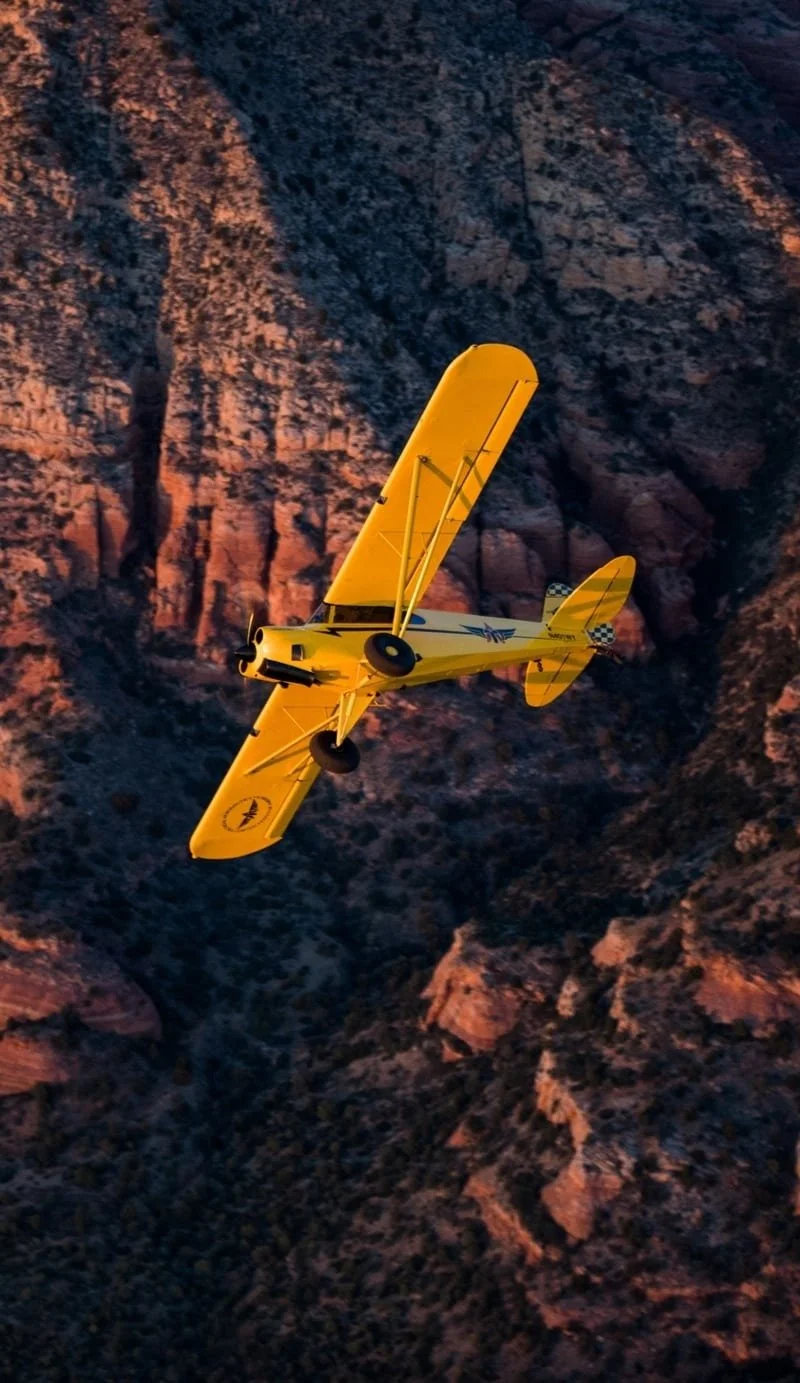 A yellow biplane flying over mountainous terrain during sunset.