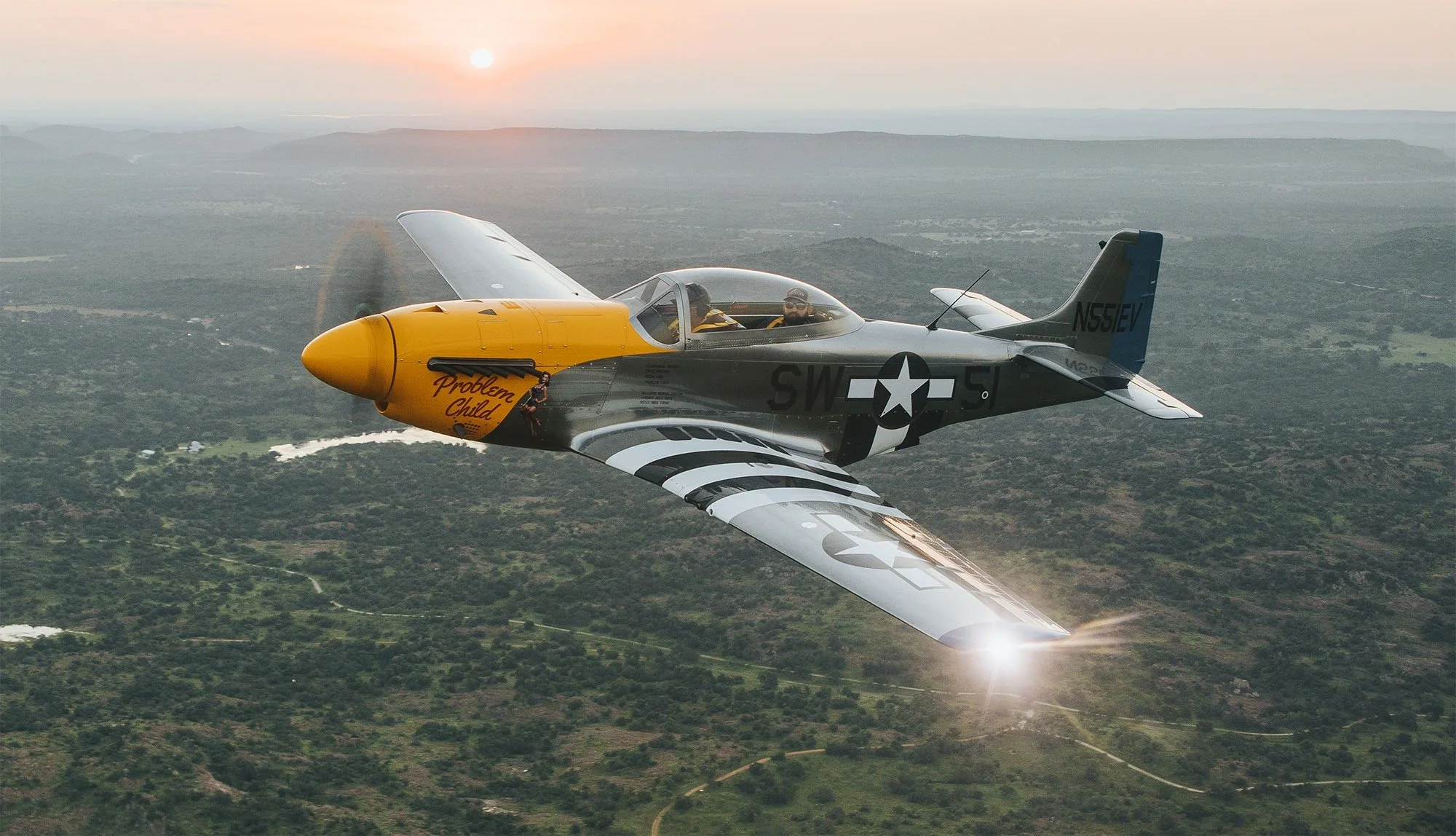 A vintage fighter jet painted with a yellow nose and black, gray, and white body, flying over a landscape during sunset.