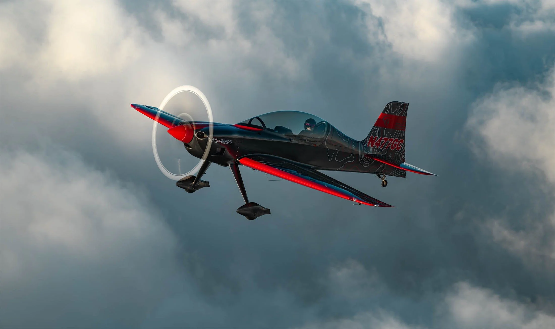 A small black and red aerobatic airplane flying through cloudy sky, with a pilot visible inside the cockpit.
