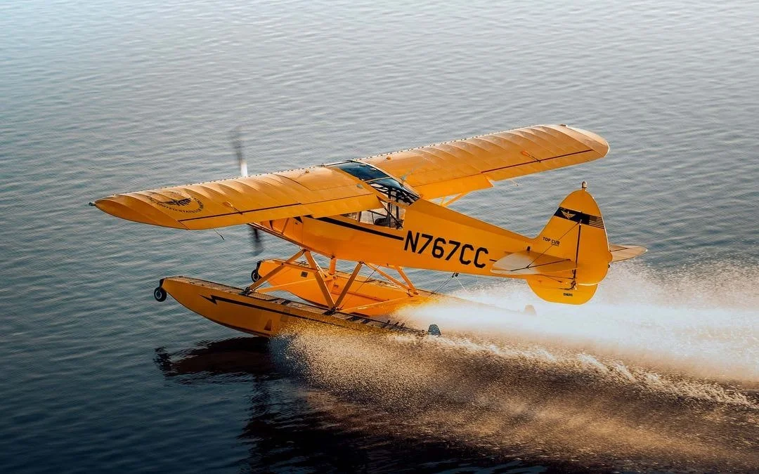 A yellow seaplane flying low over water, creating a spray behind it, registration N767CC