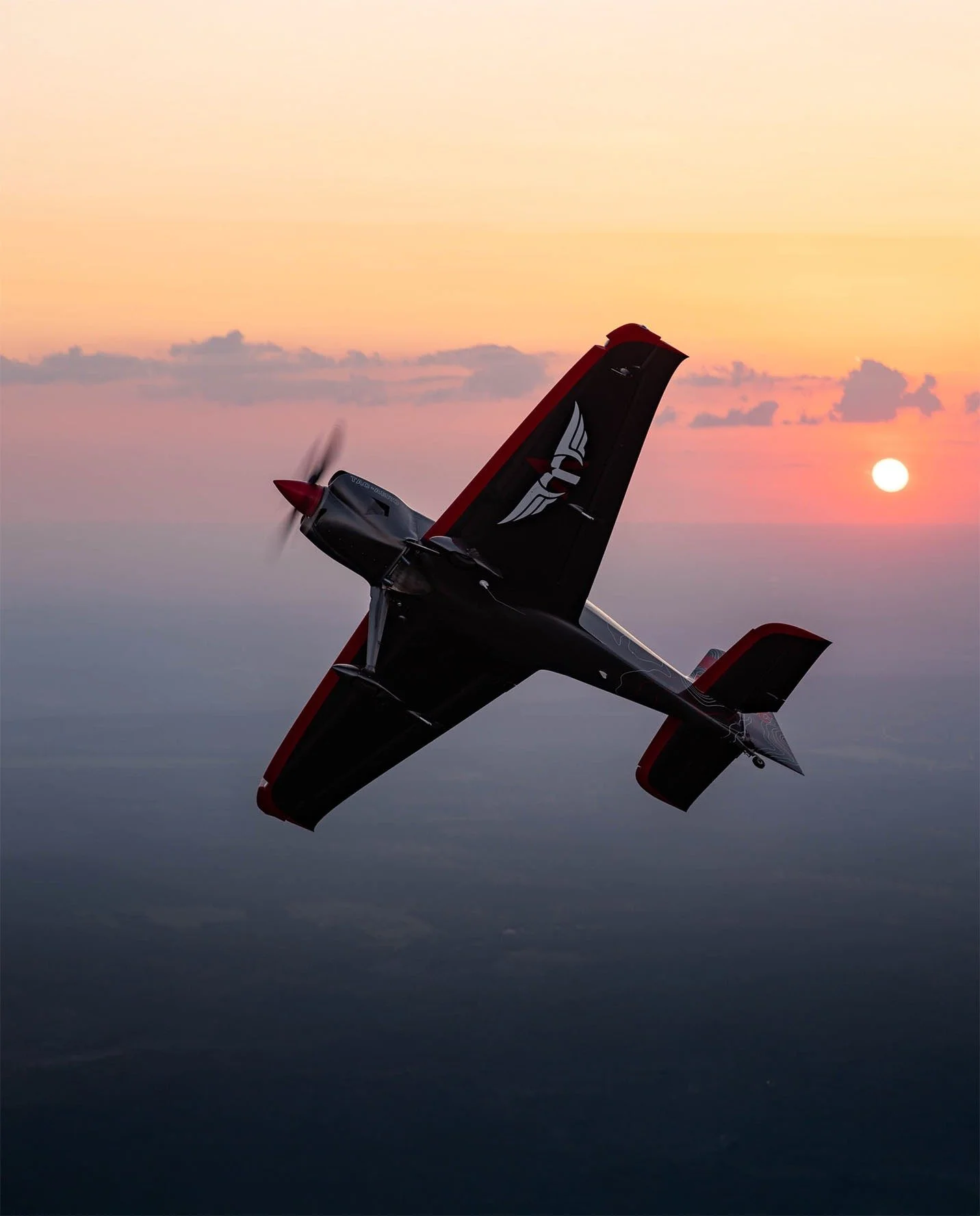 A black and red aerobatic airplane flying during sunset with a sunset in the background.