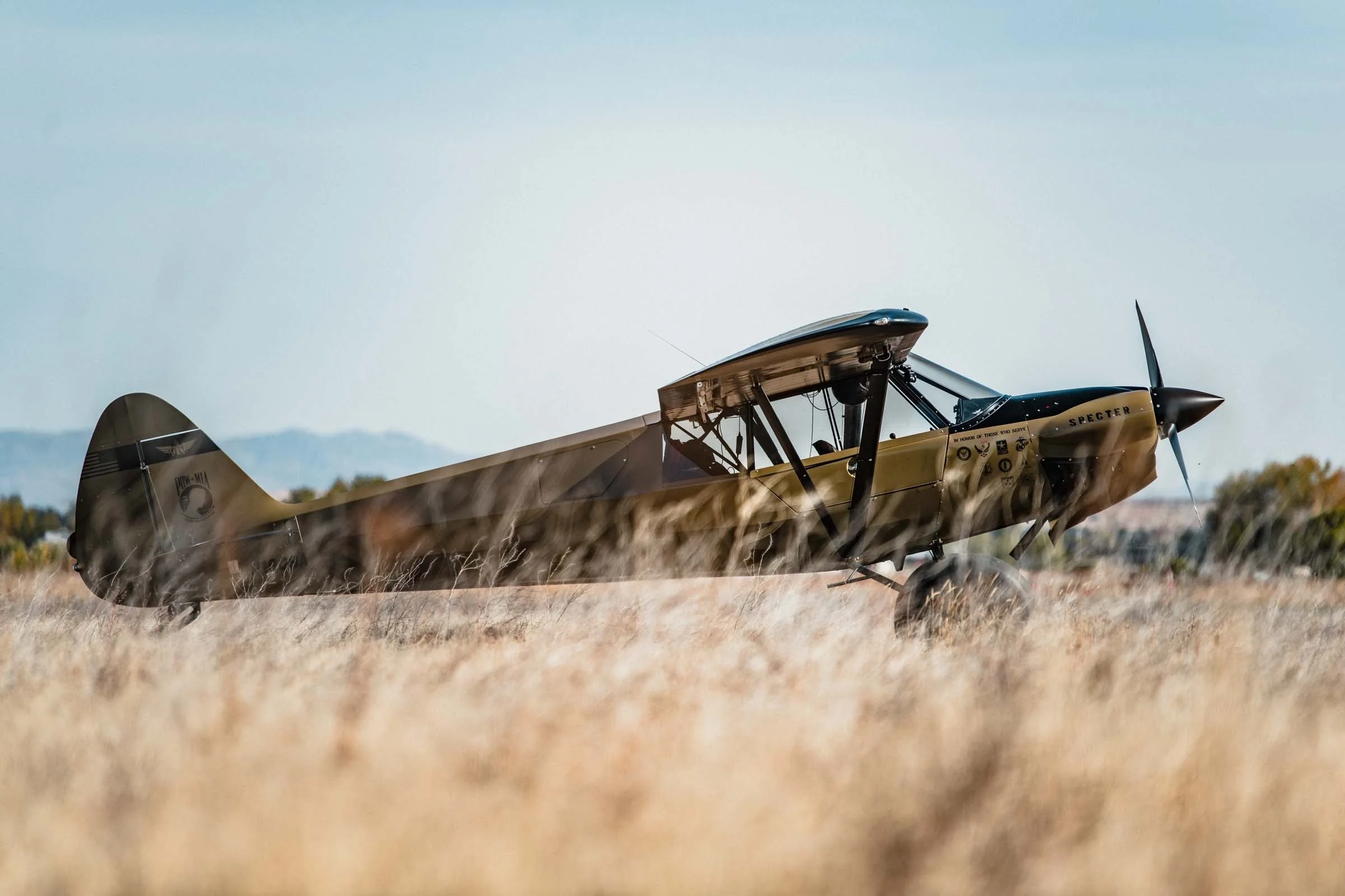 A small vintage aircraft, painted in tan and black, is parked on a grassy field with mountains in the background.
