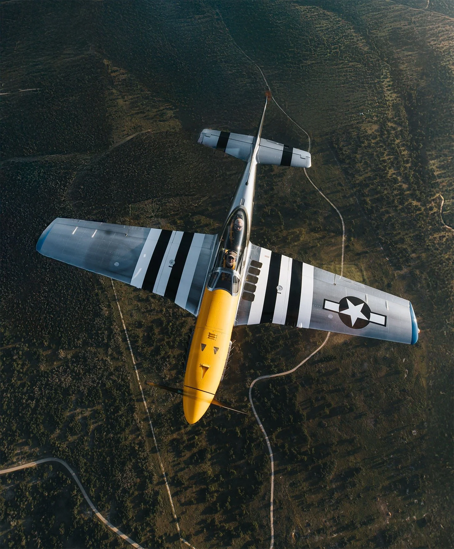 An aerial view of a military aircraft flying over a forested landscape with winding roads.