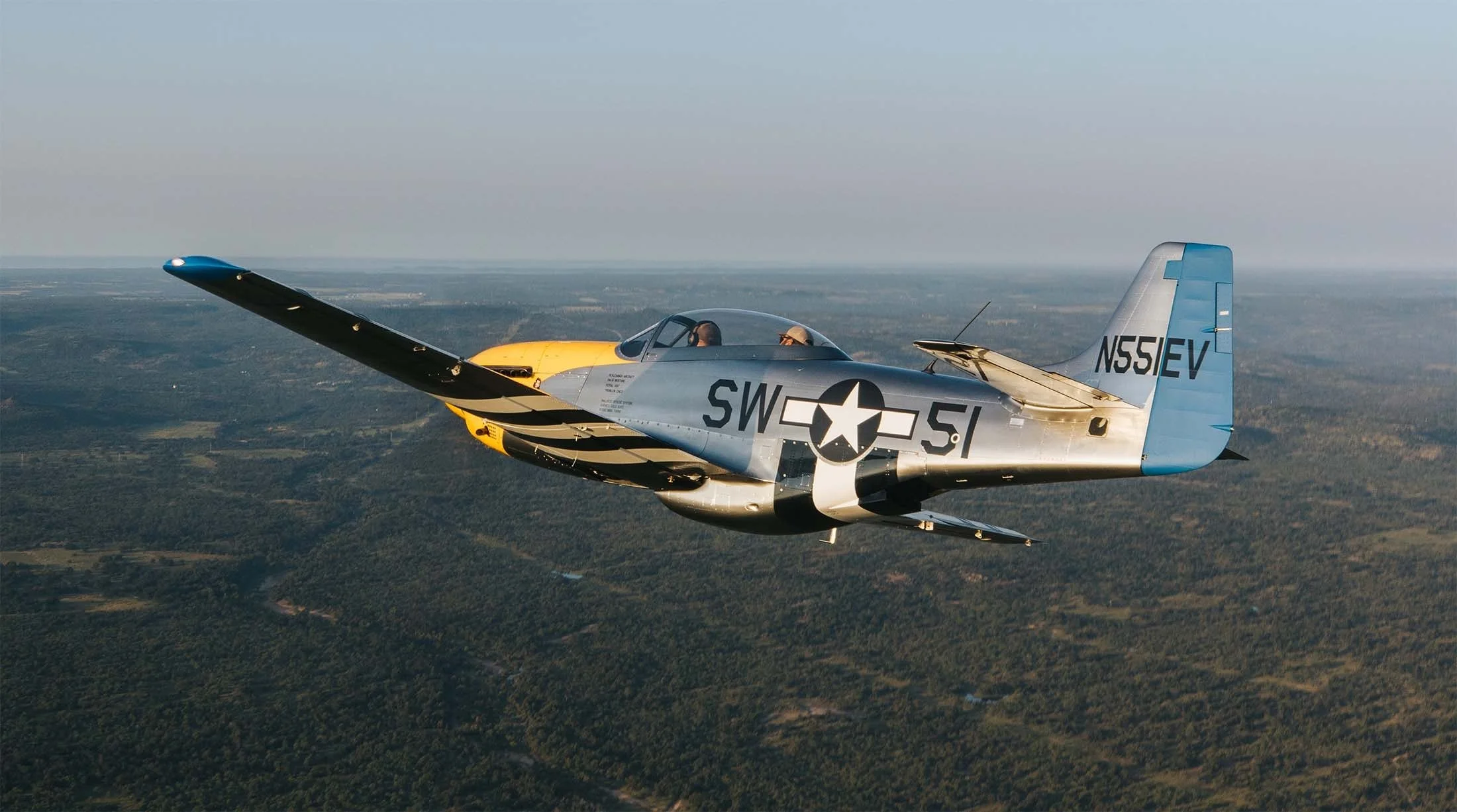 A silver and yellow vintage military jet airplane flying over a landscape with trees and fields.