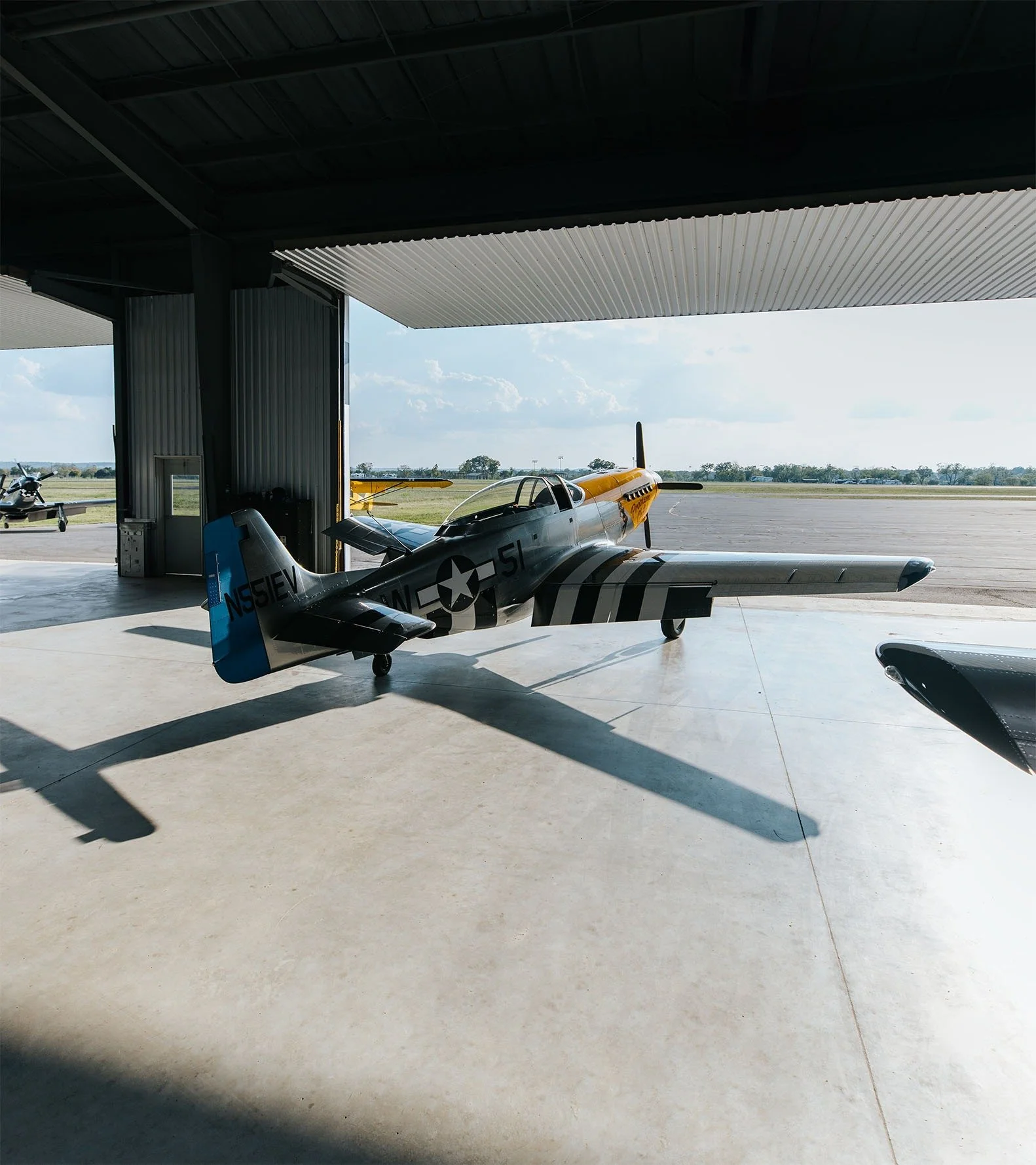 Vintage military aircraft with camouflage paint inside a hangar, with a view of the runway outside.