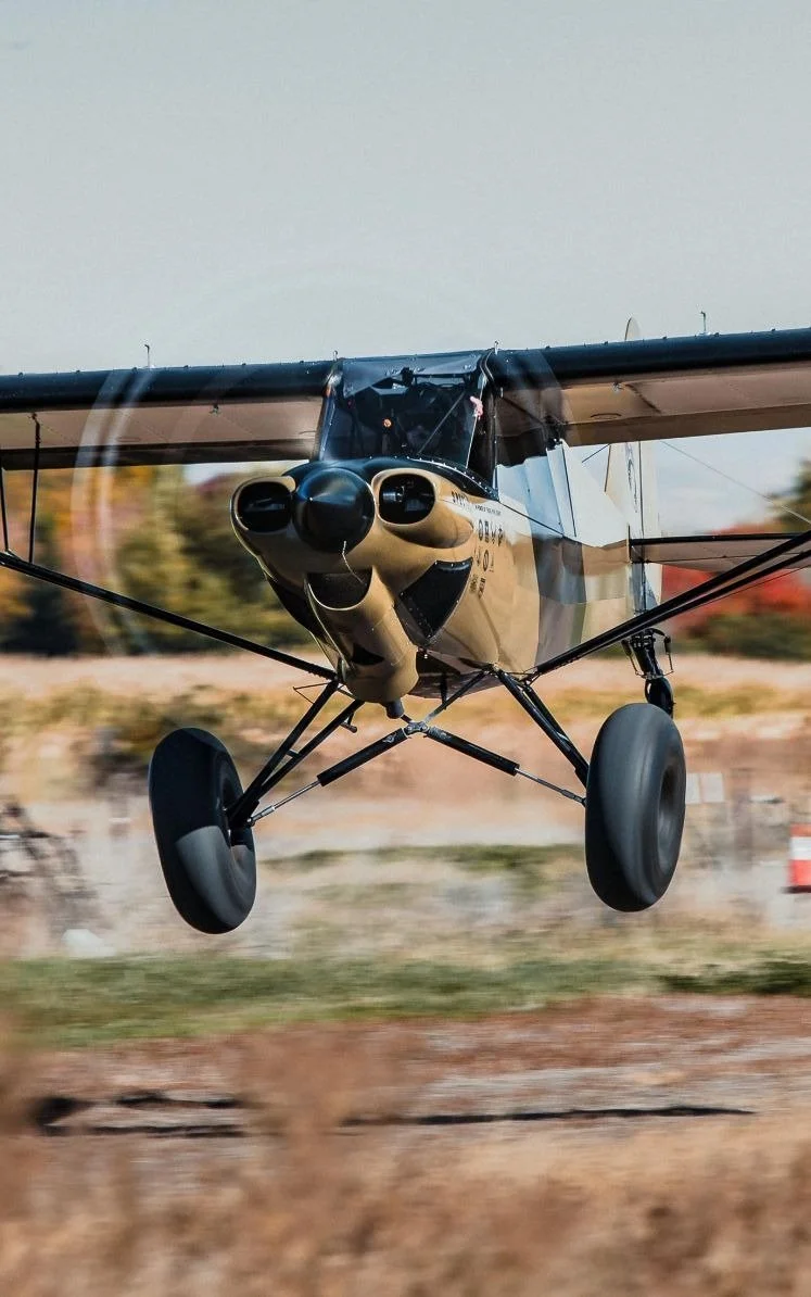 Small beige and black propeller airplane flying low over a runway with a blurred background of trees and construction cones.