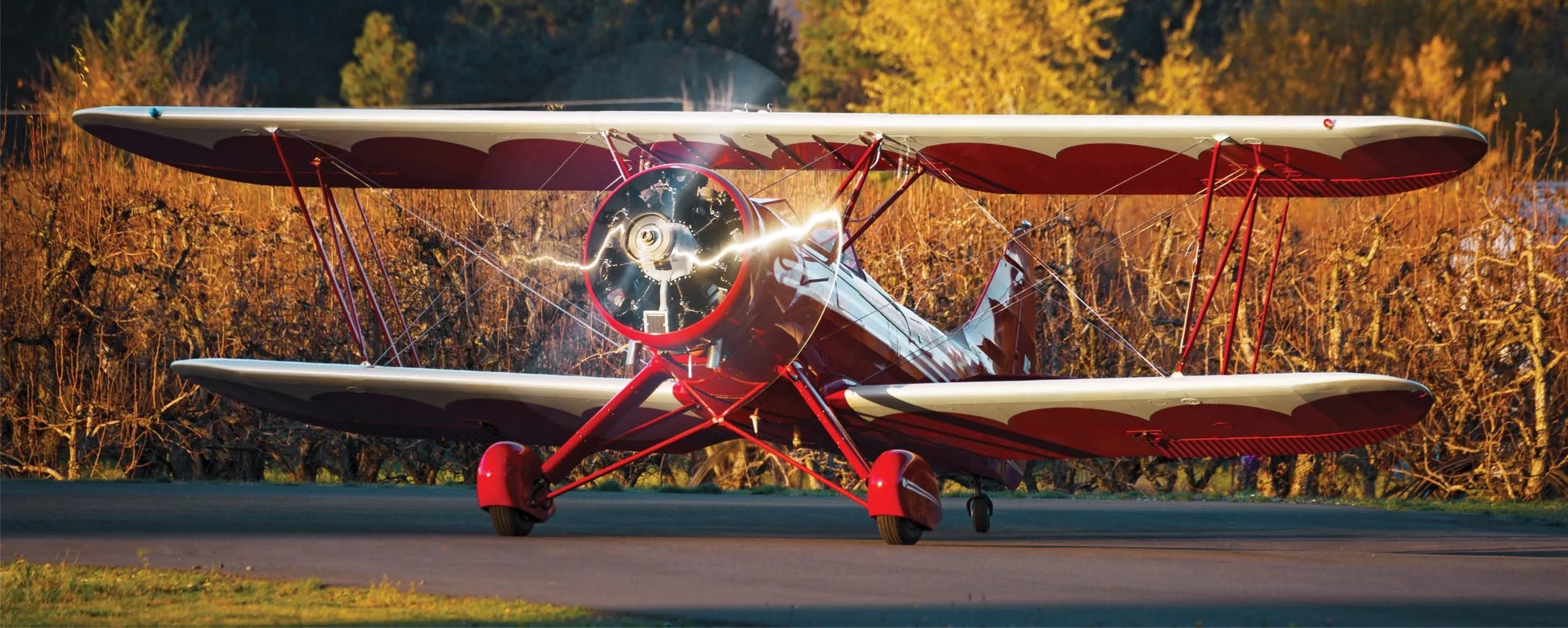 A vintage red and white biplane on a runway with the propeller spinning, surrounded by autumn trees with orange and yellow leaves.