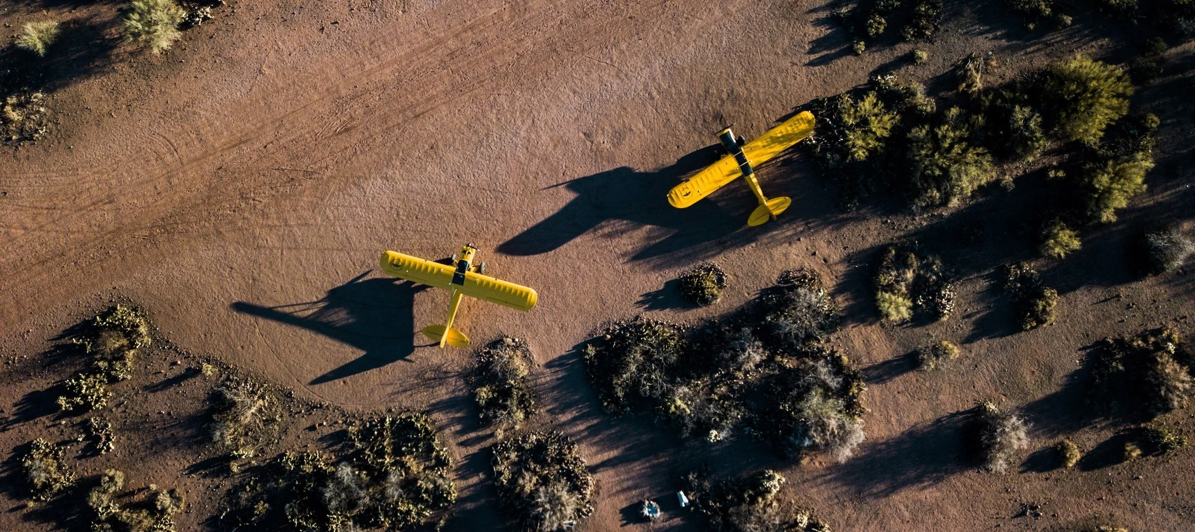 Two yellow crop duster airplanes flying over a desert landscape with sparse vegetation and shrubs, casting long shadows.